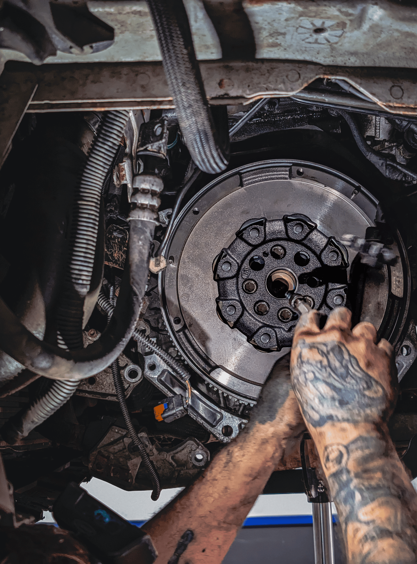 A mechanic with tattooed arms repairs a car engine flywheel and clutch assembly in an auto shop.