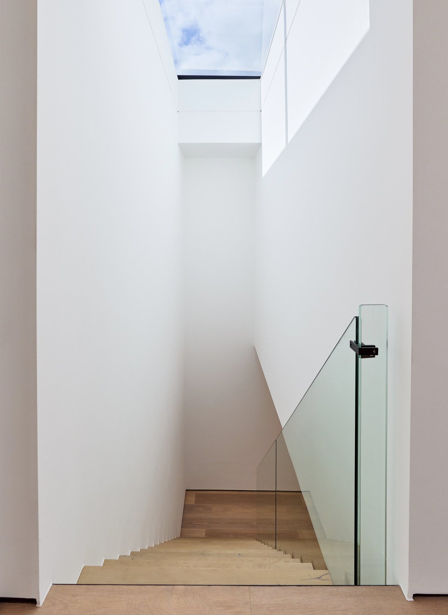 Modern des staircase with glass railing leading to a bright skylight in a minimalist white interior.