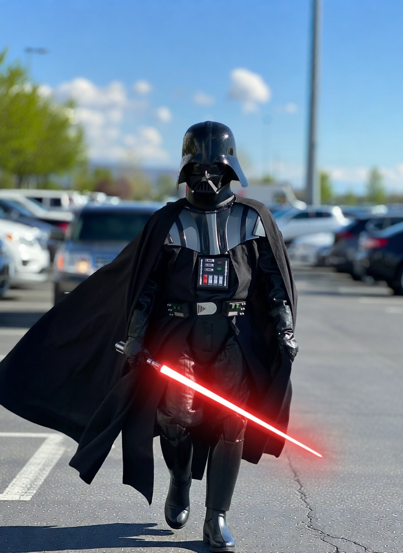 A person in a realistic Darth Vader cosplay costume holding a glowing red lightsaber in a sunny parking lot.