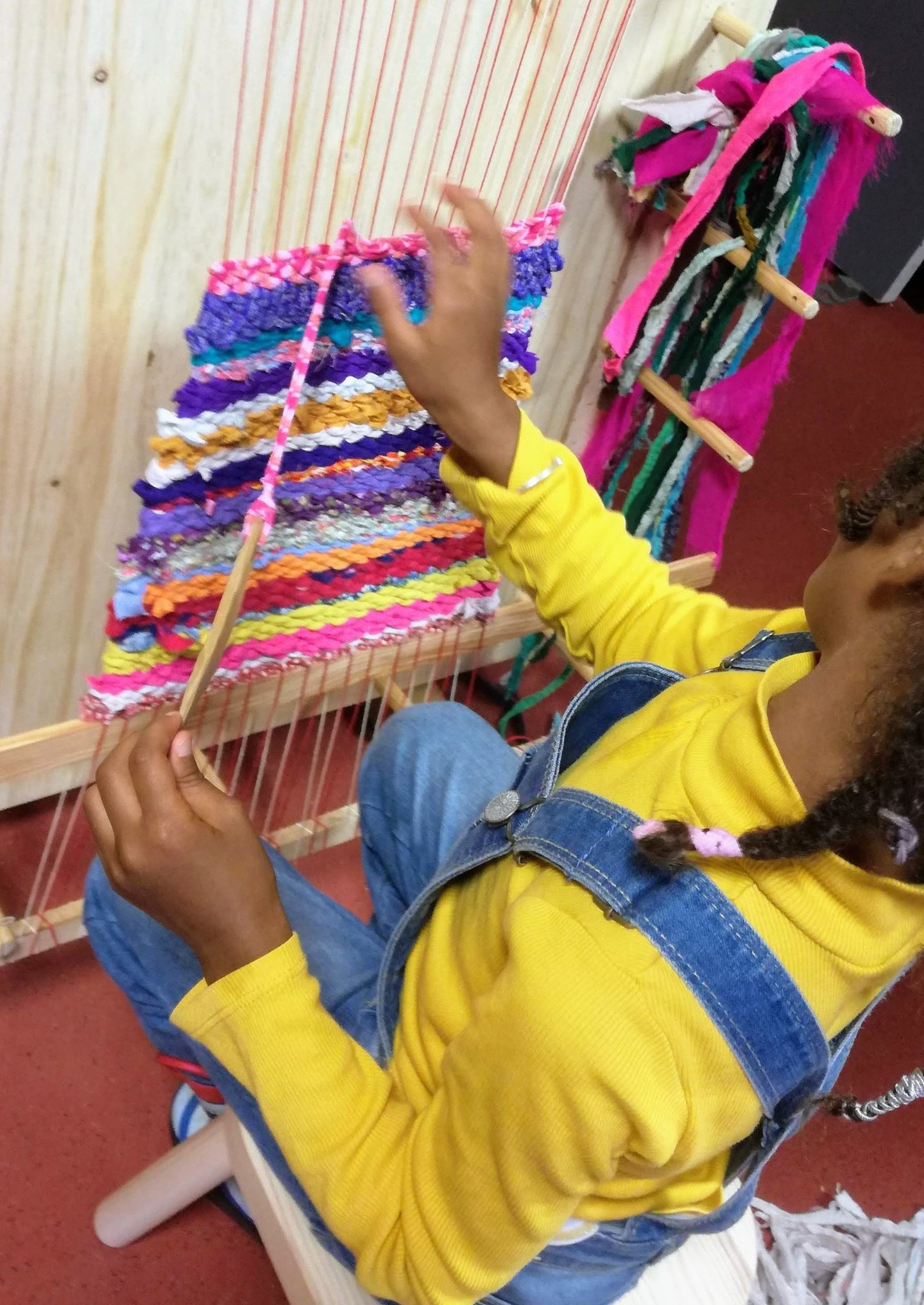 a young girl sits on a chair and weaves colorful rag rug on a weaving loom