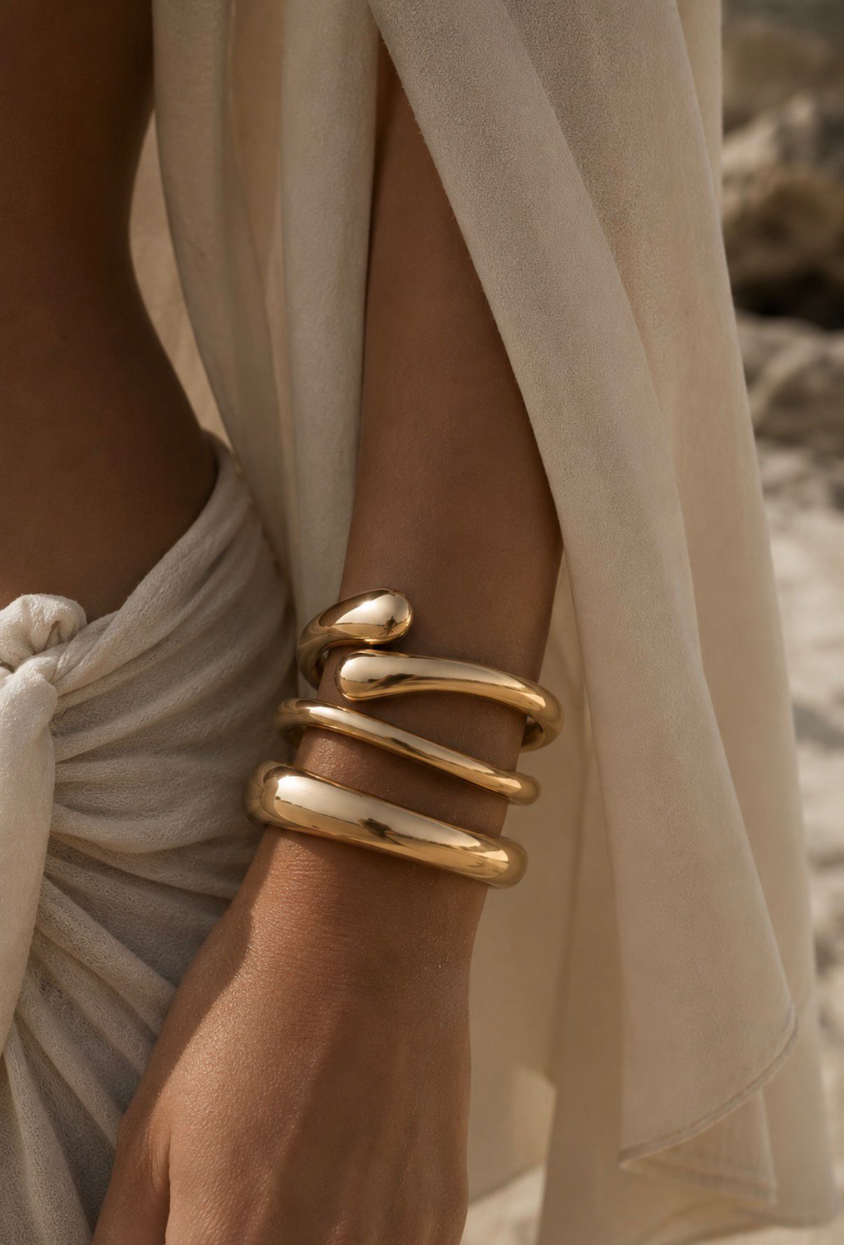 Gold statement bracelets styled on an arm with a beige beach cover-up in warm sunlight.