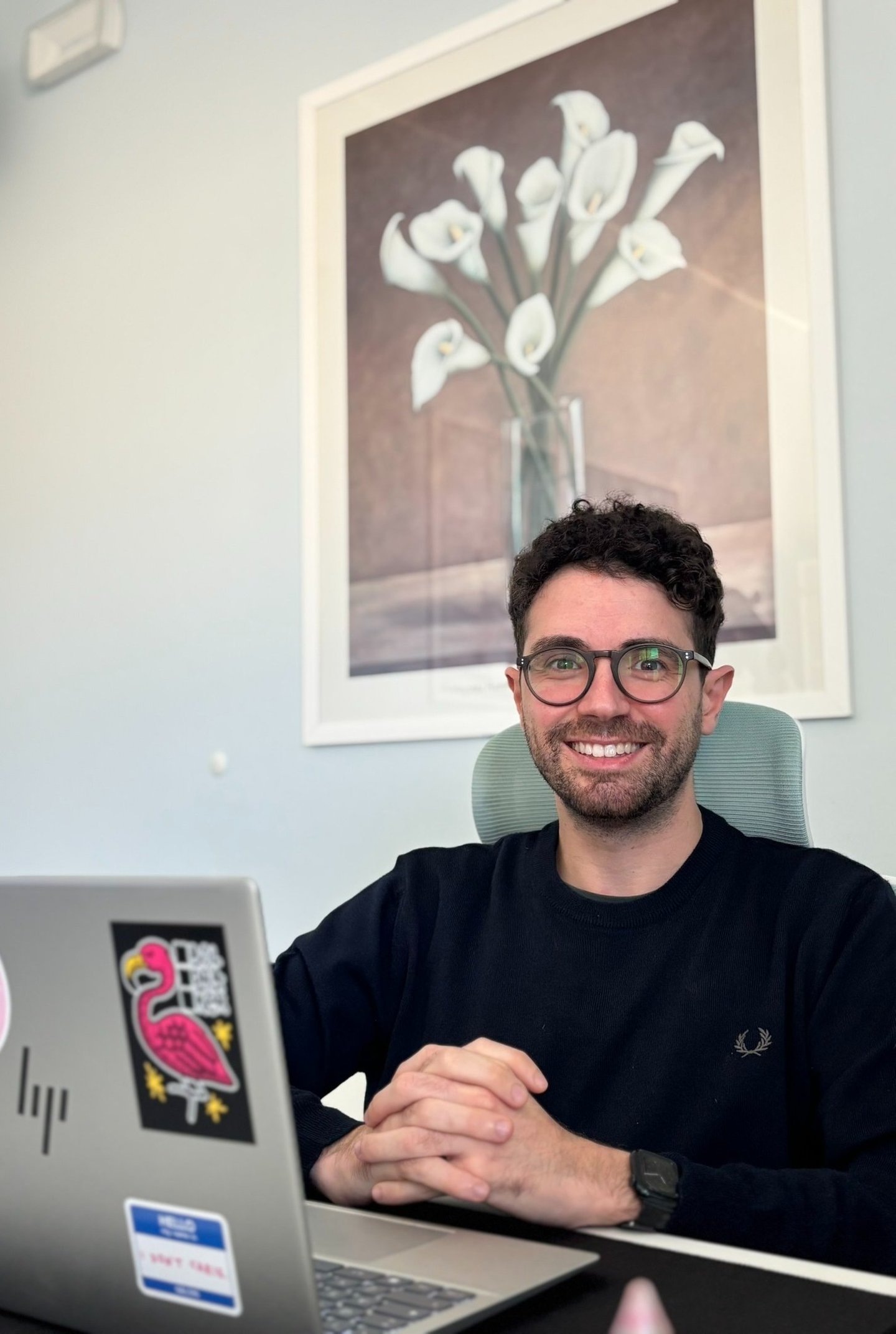 Smiling man with glasses sitting at a desk with a laptop in a modern office setting.