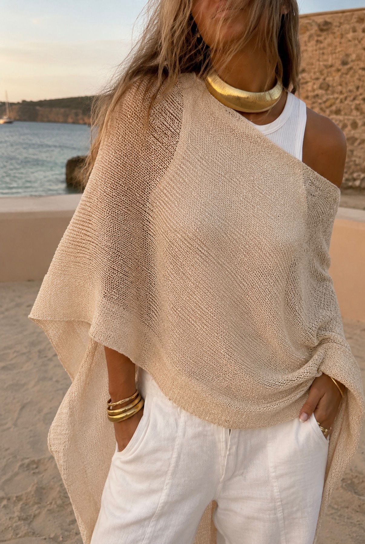 Woman wearing a beige knit cape top cover-up over a white tank top with gold statement jewelry.