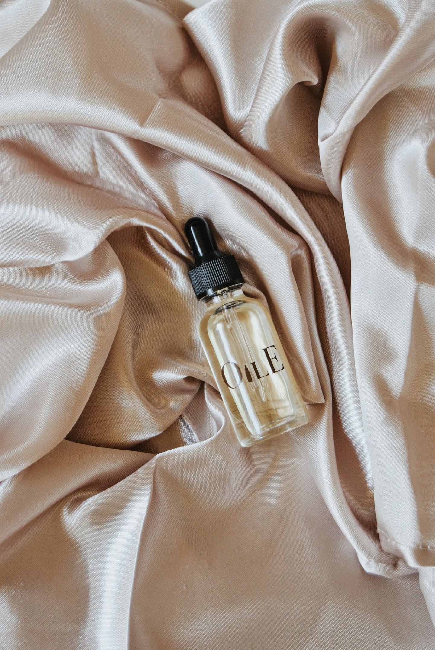 A glass facial oil dropper bottle resting on luxurious champagne silk fabric.
