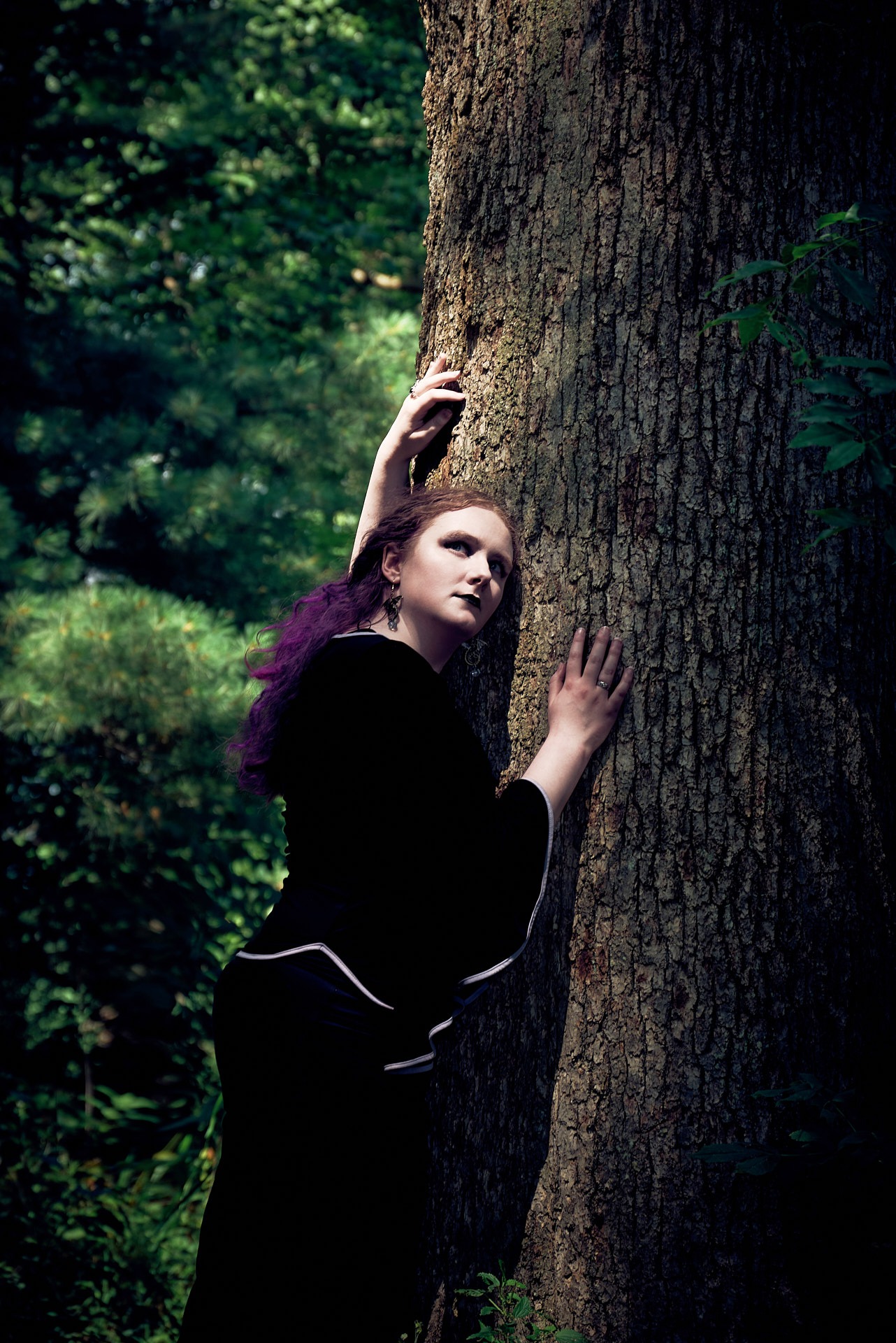 a woman with purple hair in a black dress in the forest with sunlight on her face