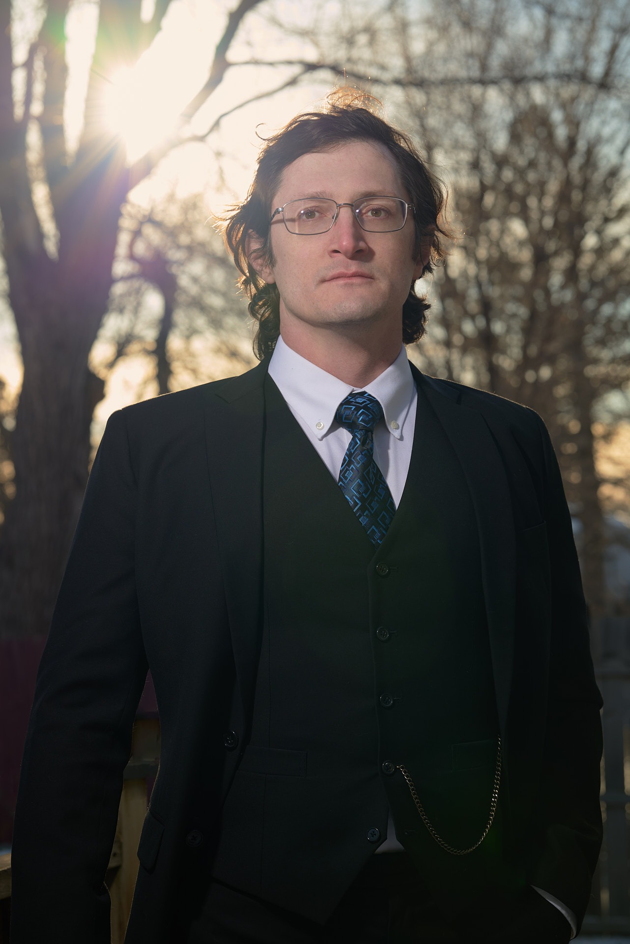 a man in a  black suit and glasses and tie with a blue tie in front of the sun and trees