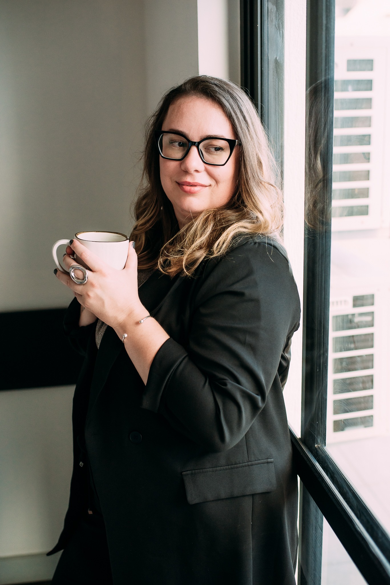 Uma mulher profissional vestindo um blazer preto, segurando uma caneca de café perto de uma janela i