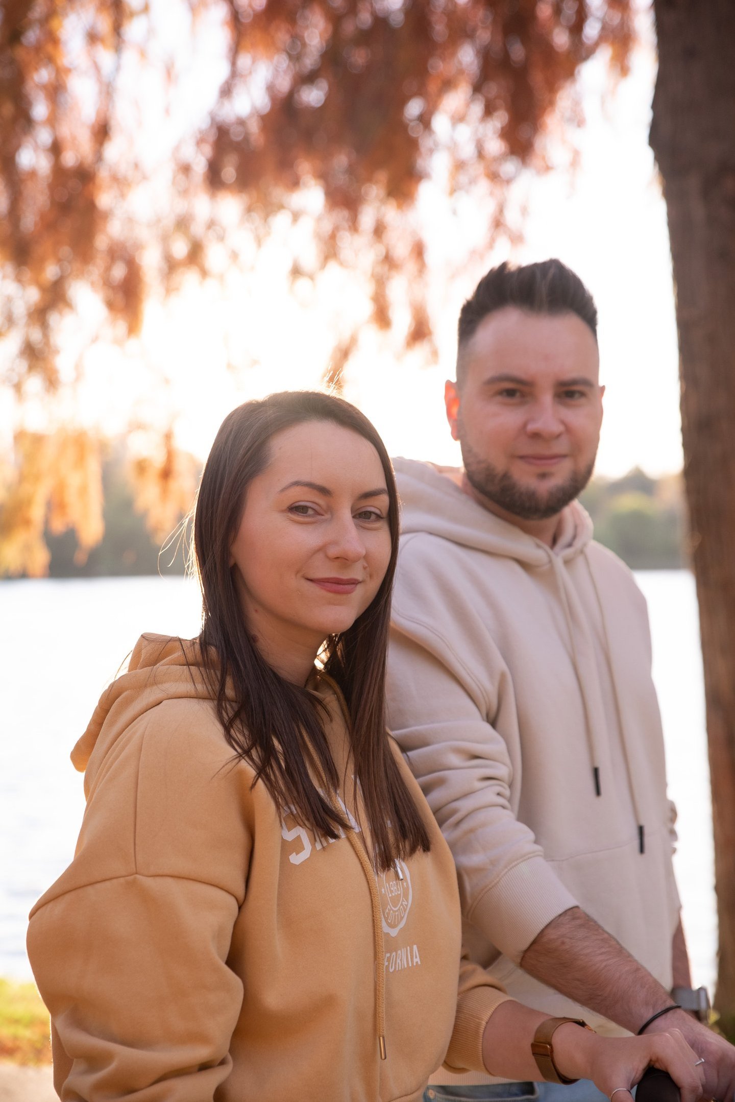Couple wearing hoodies standing outdoors by a lake under autumn foliage at golden hour.