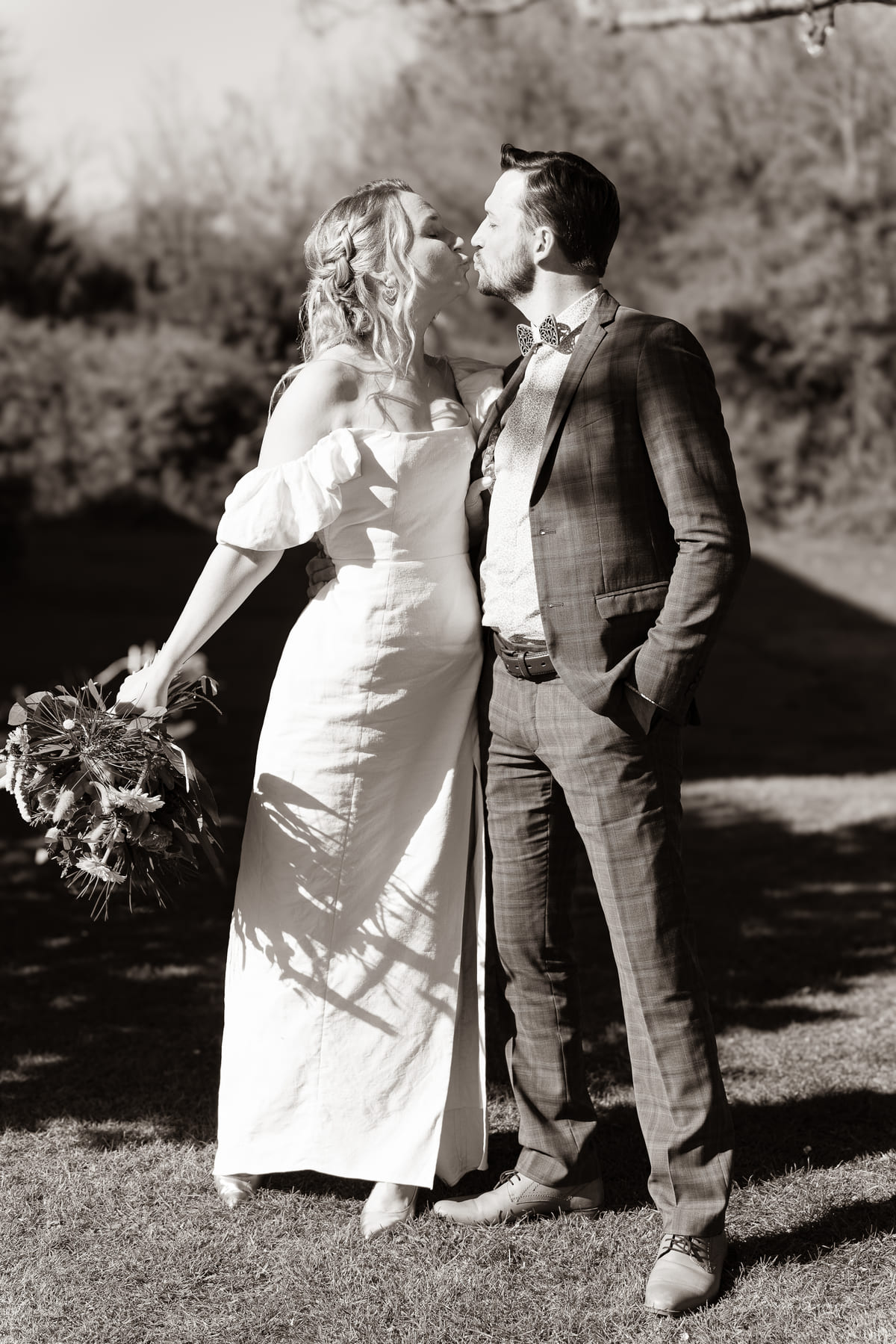 A bride in a white wedding dress and a groom in a suit sharing a kiss outdoors.