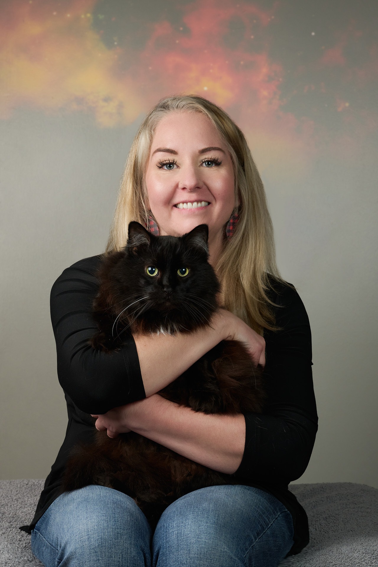 A smiling blonde woman holding a fluffy black long-haired cat in a studio portrait with a galaxy background.