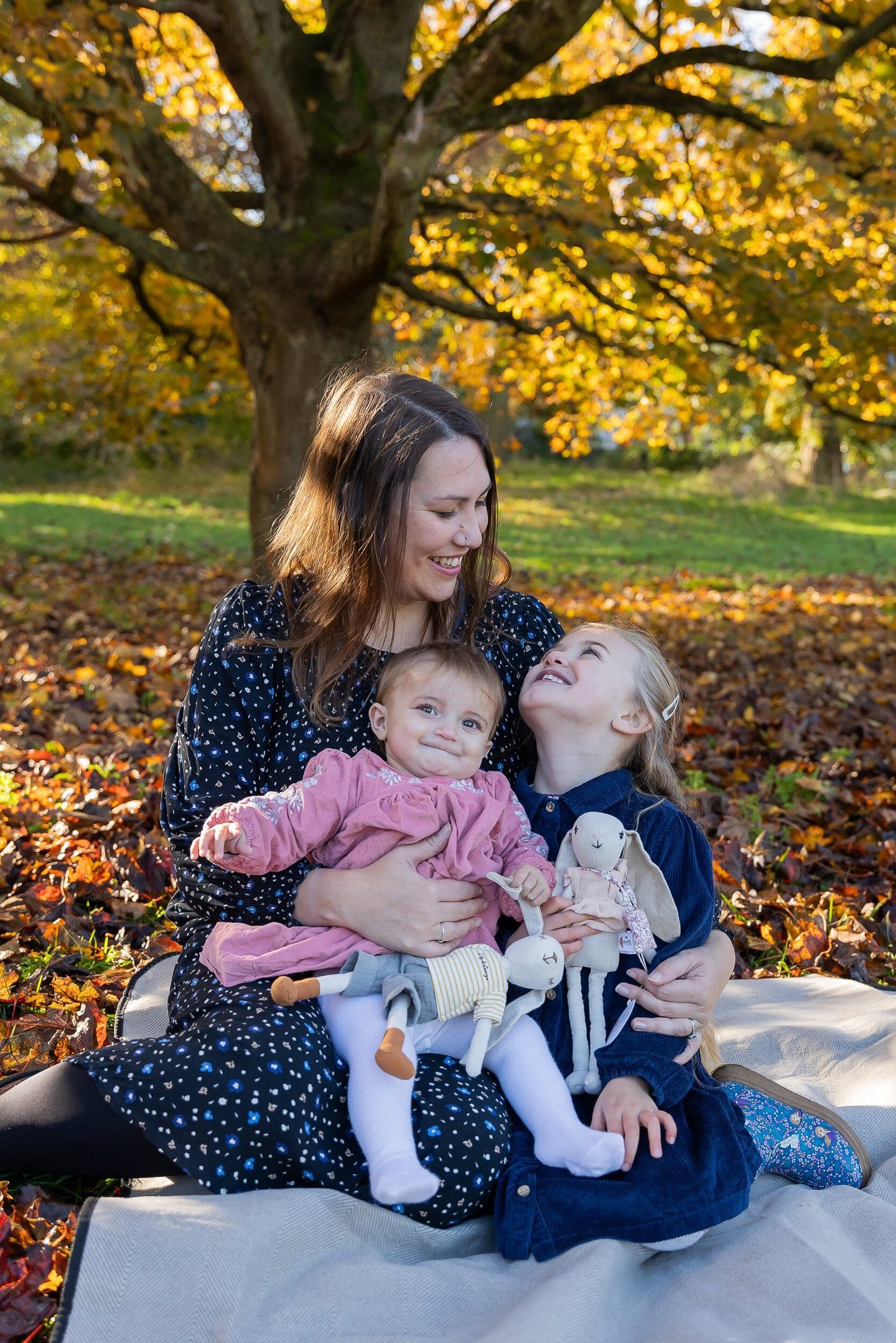 A mother and two daughters sitting on fall leaves under a golden autumn tree for a family photoshoot.