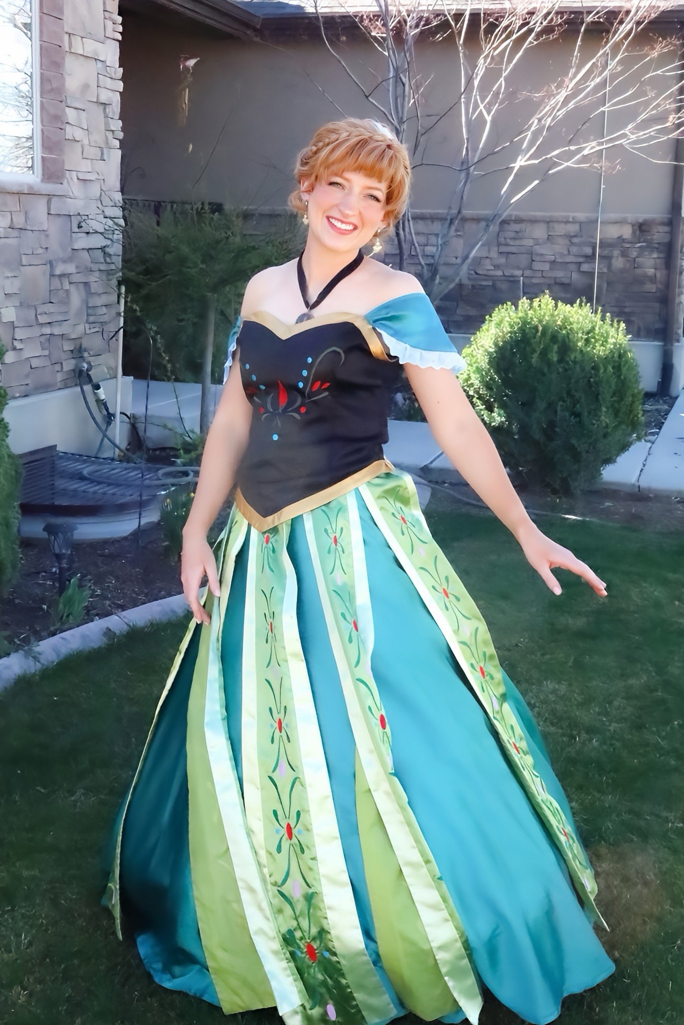 Smiling woman in a detailed Princess Anna coronation dress cosplay standing in a garden.