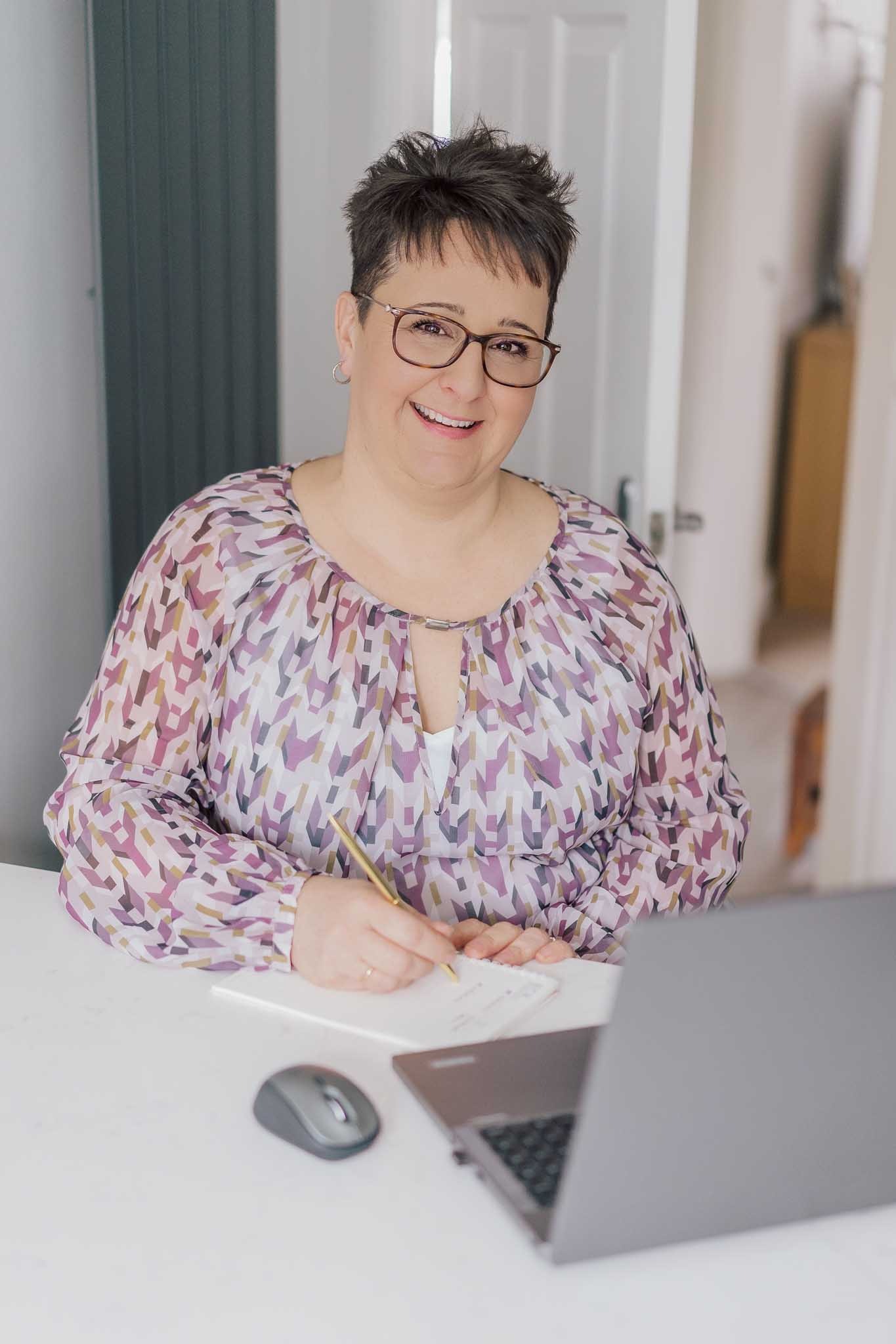 A smiling professional woman with glasses taking notes at her desk with a laptop.
