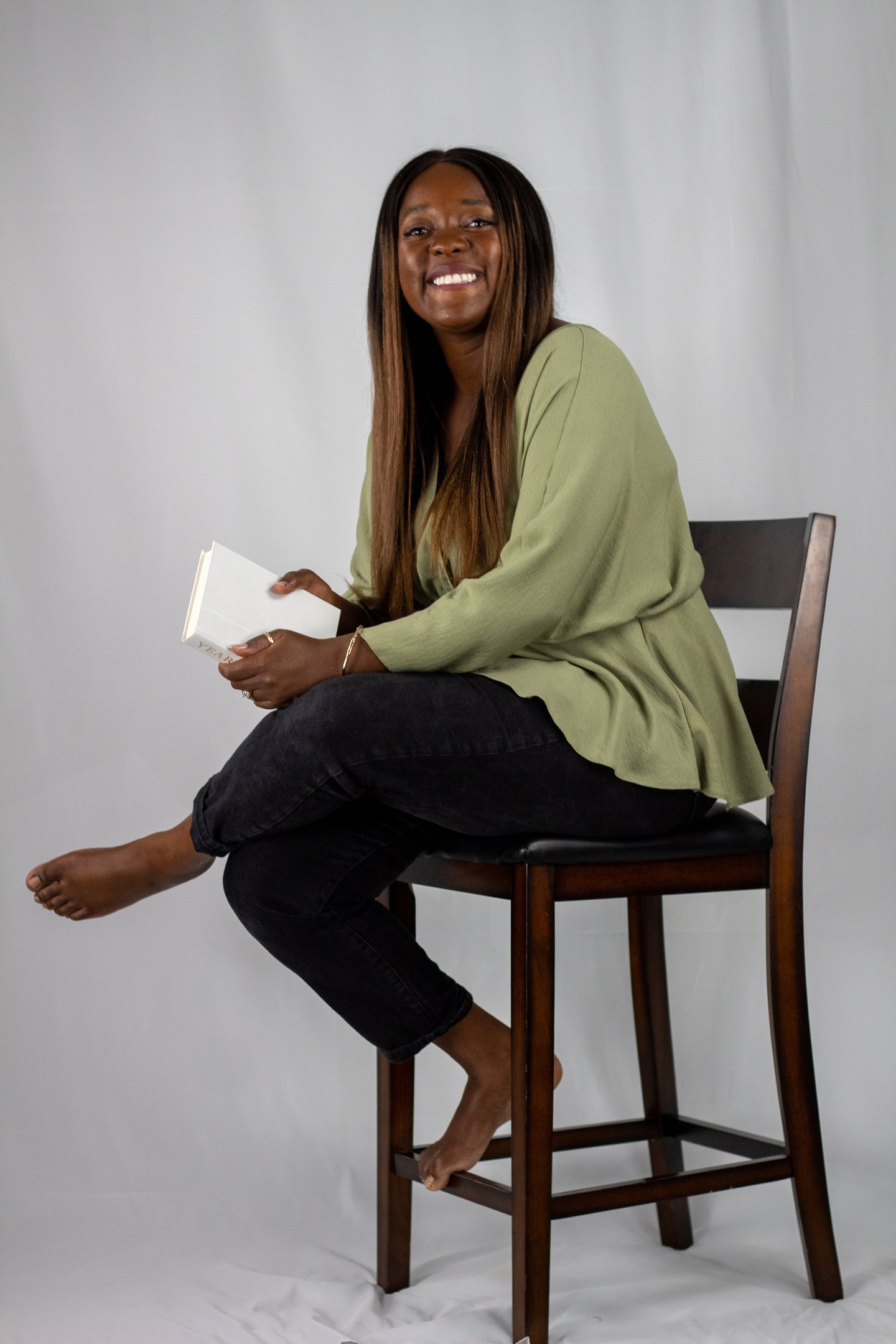 Esther Osaheni, founder of Incurable Optimist, seated on a wooden chair holding a book and smiling
