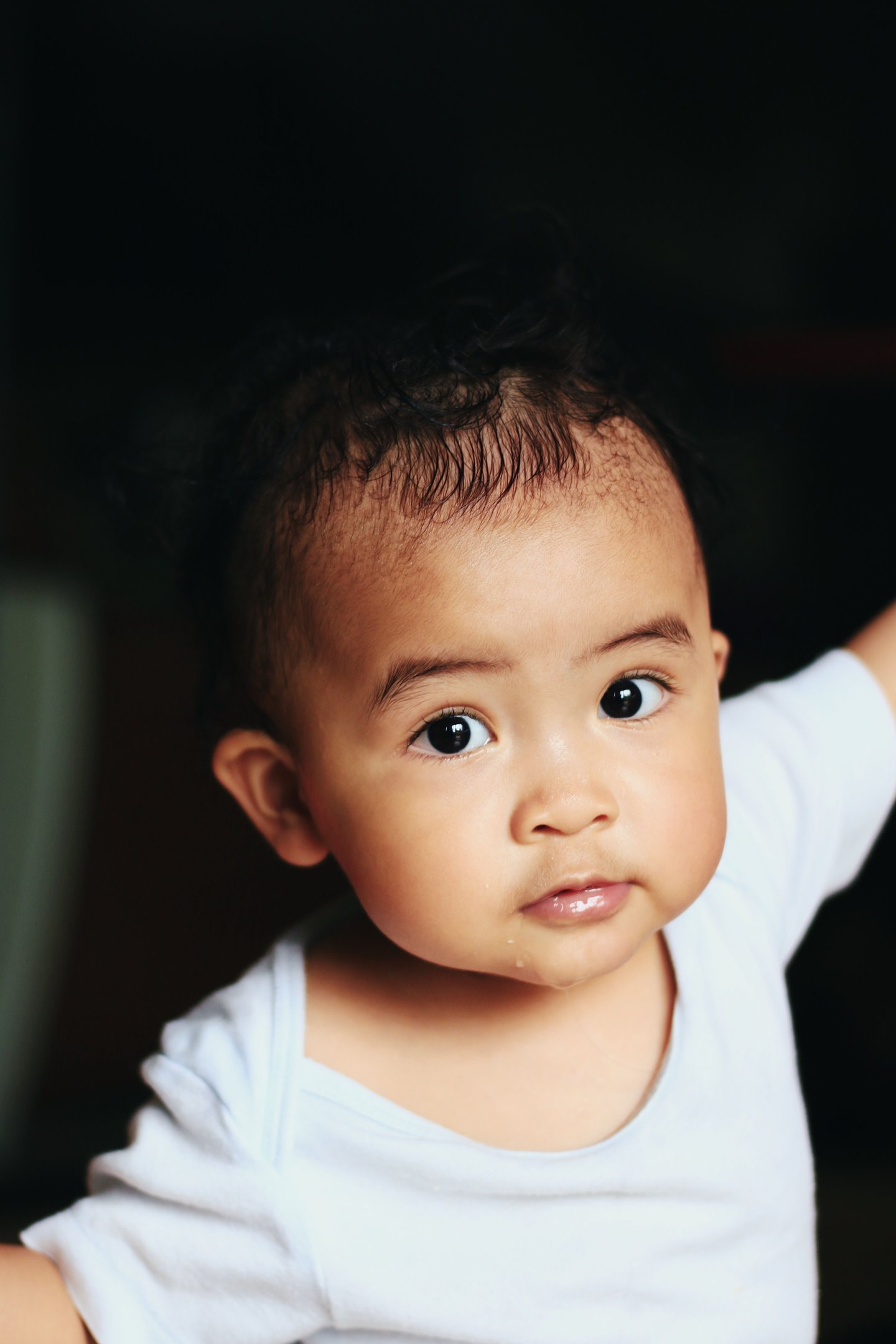 A close-up portrait of a cute baby with dark curly hair and large expressive eyes wearing a white shirt.