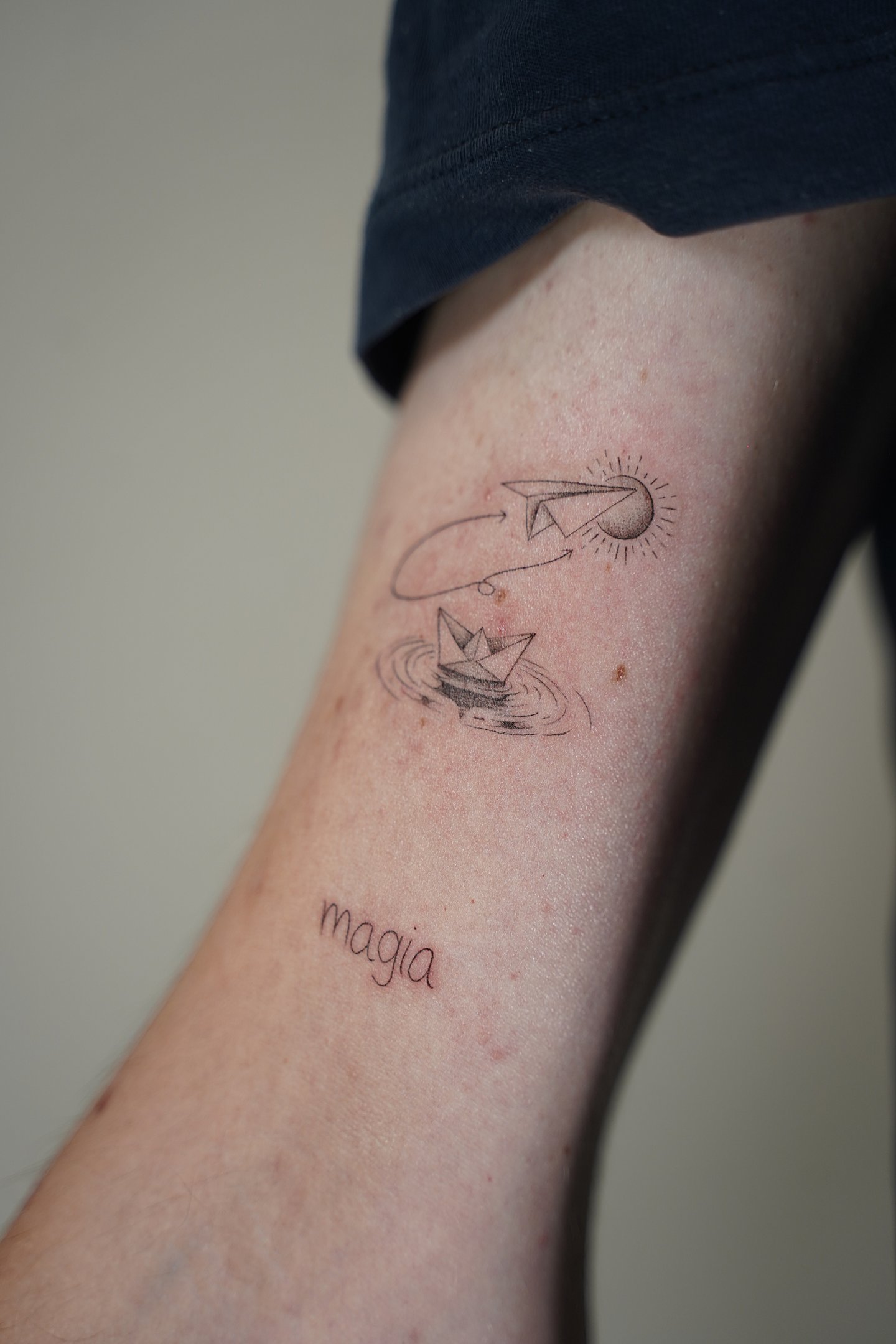 Tatuaje fineline en Madrid de barco de papel y avión de origami con luna y la palabra magia