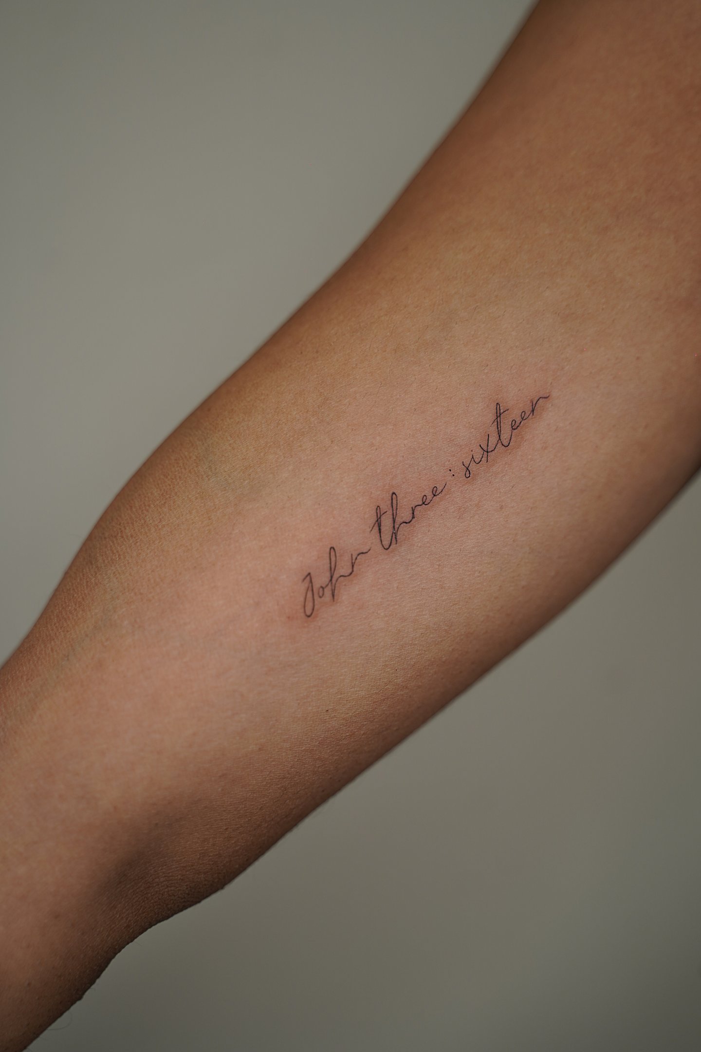 Tatuaje fineline en Madrid de frase John three sixteen en cursiva fina diagonal en el antebrazo