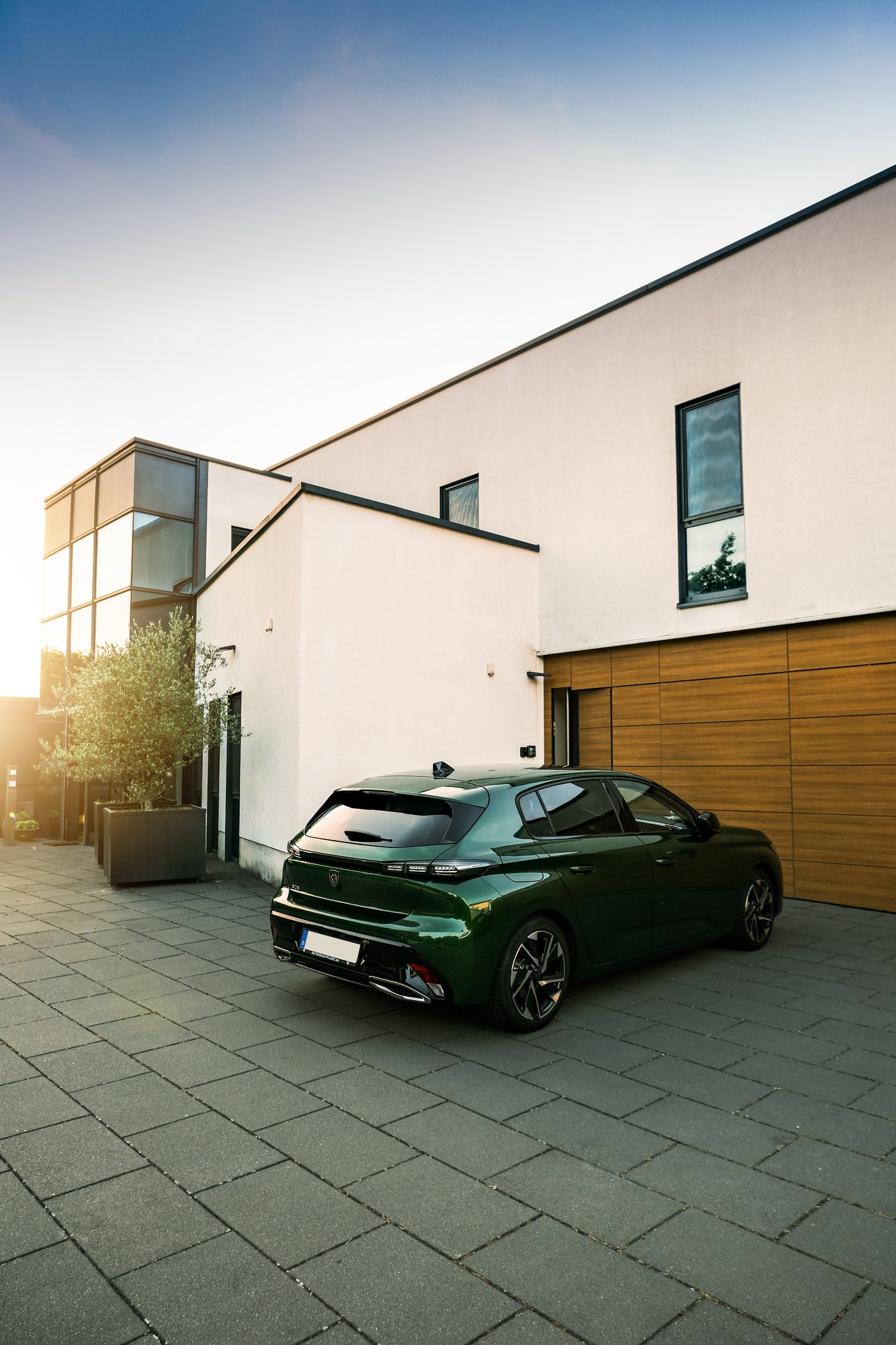 A metallic green Peugeot 308 hatchback parked on a stone driveway in front of a modern white villa.