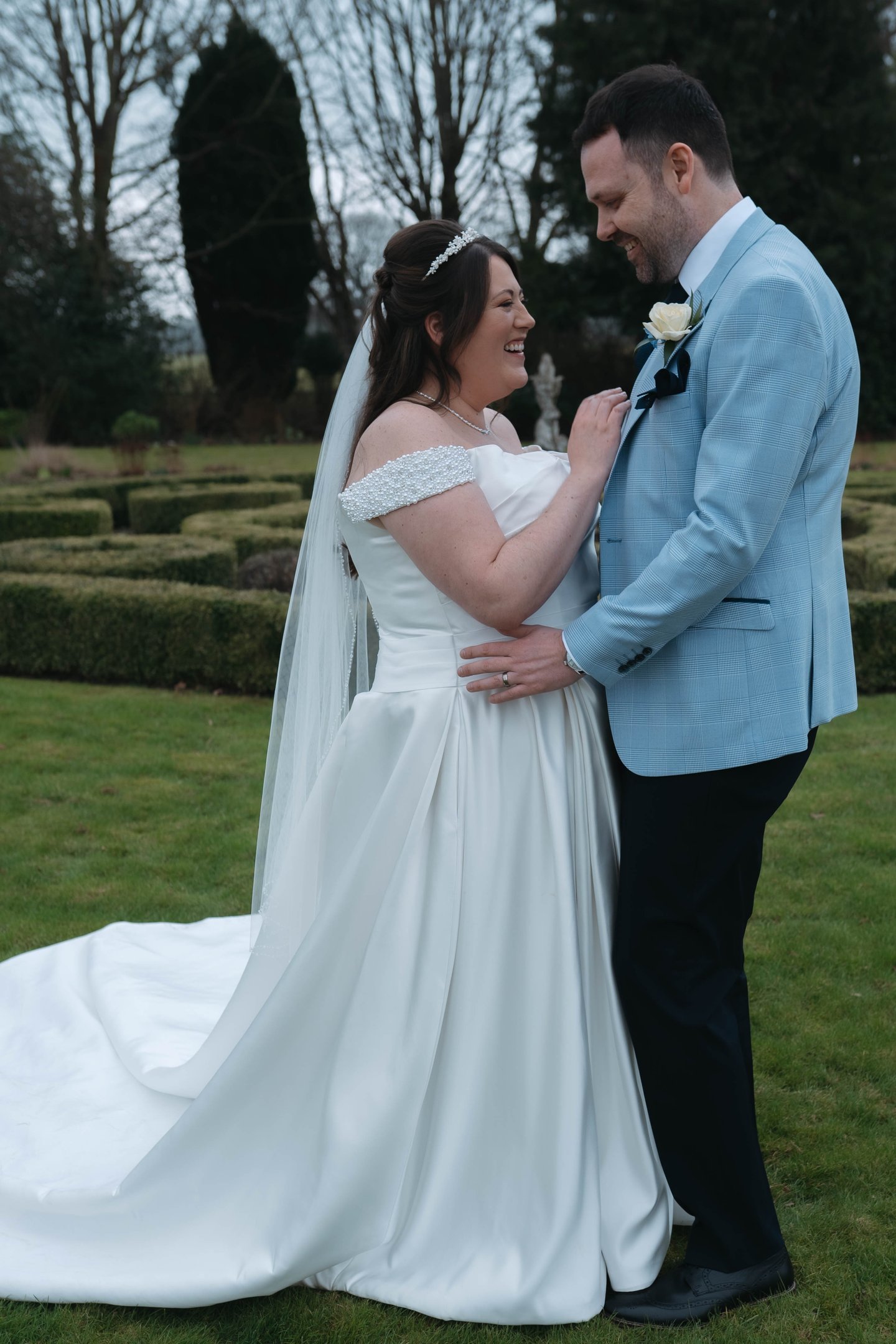A smiling bride in a white off-the-shoulder wedding dress poses with her groom in a blue suit jacket.