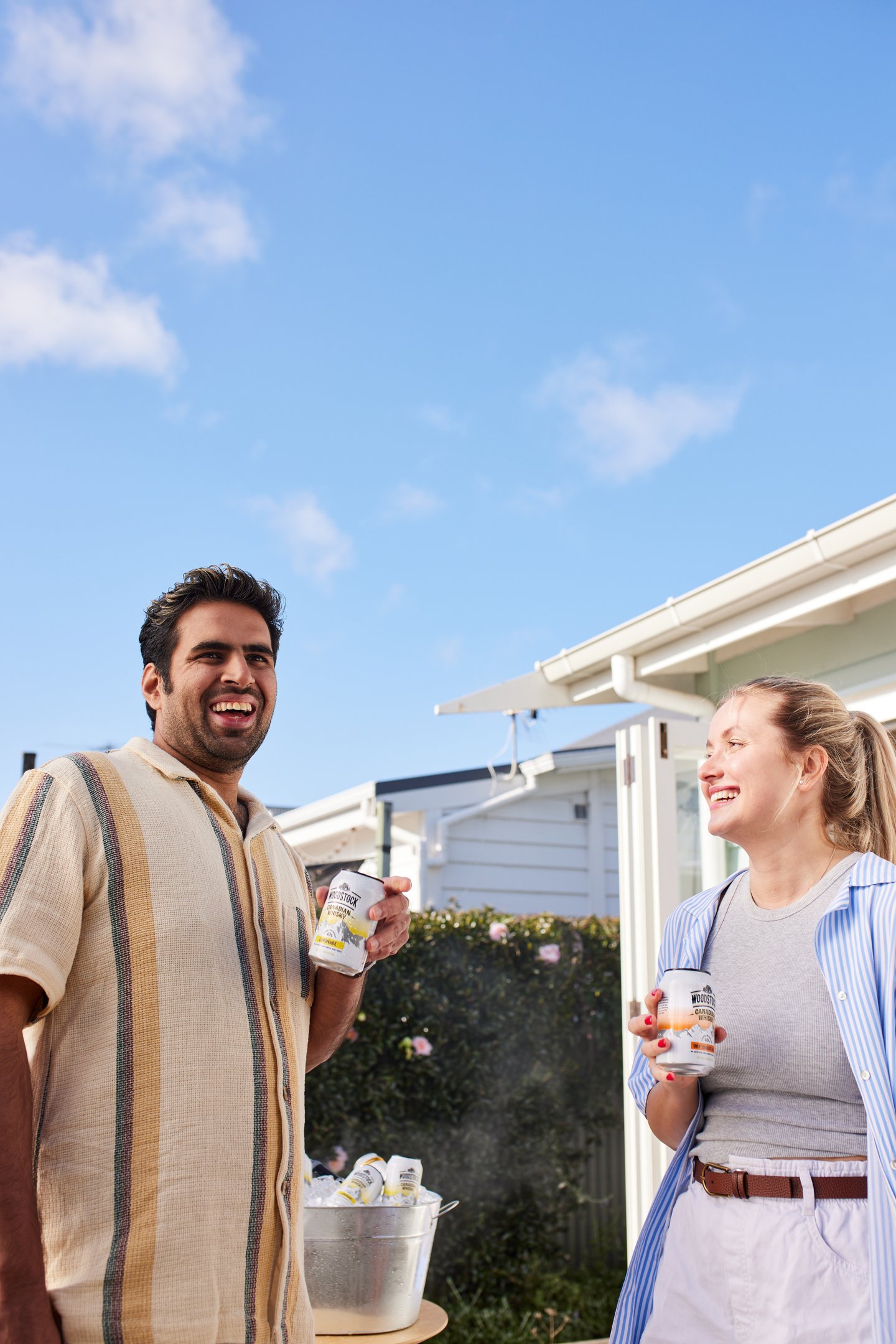 Product campaign photography - a man and woman standing outside of a house holding woodstock cans