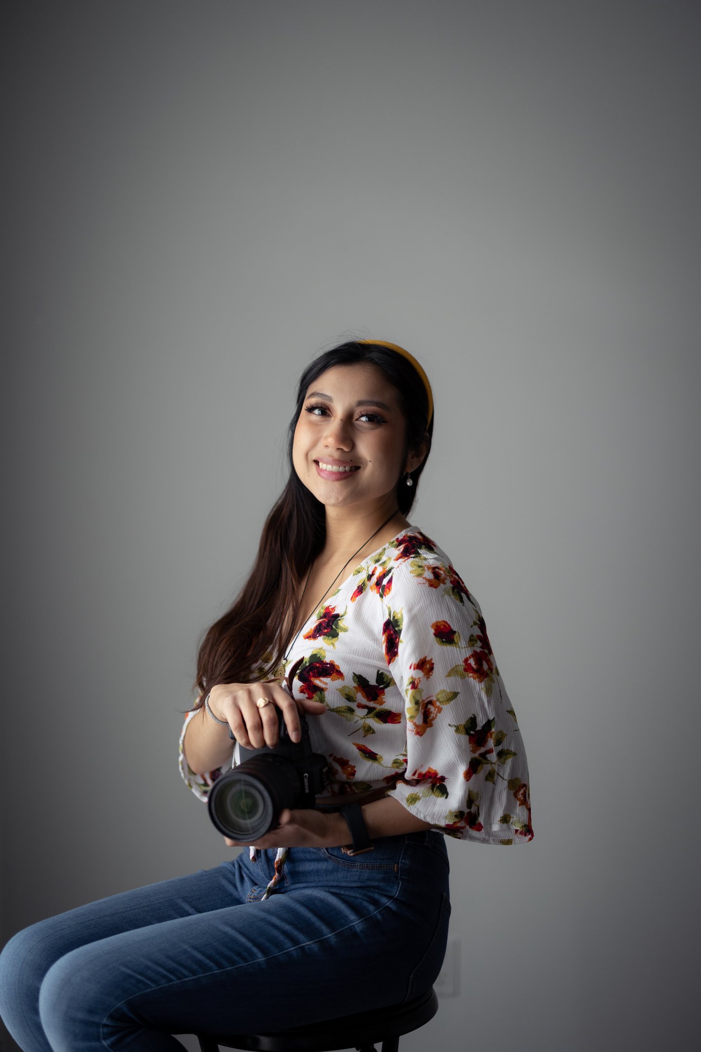 Professional shot of me holding Canon camera, sitting on a stool. Wearing floral top and headband.