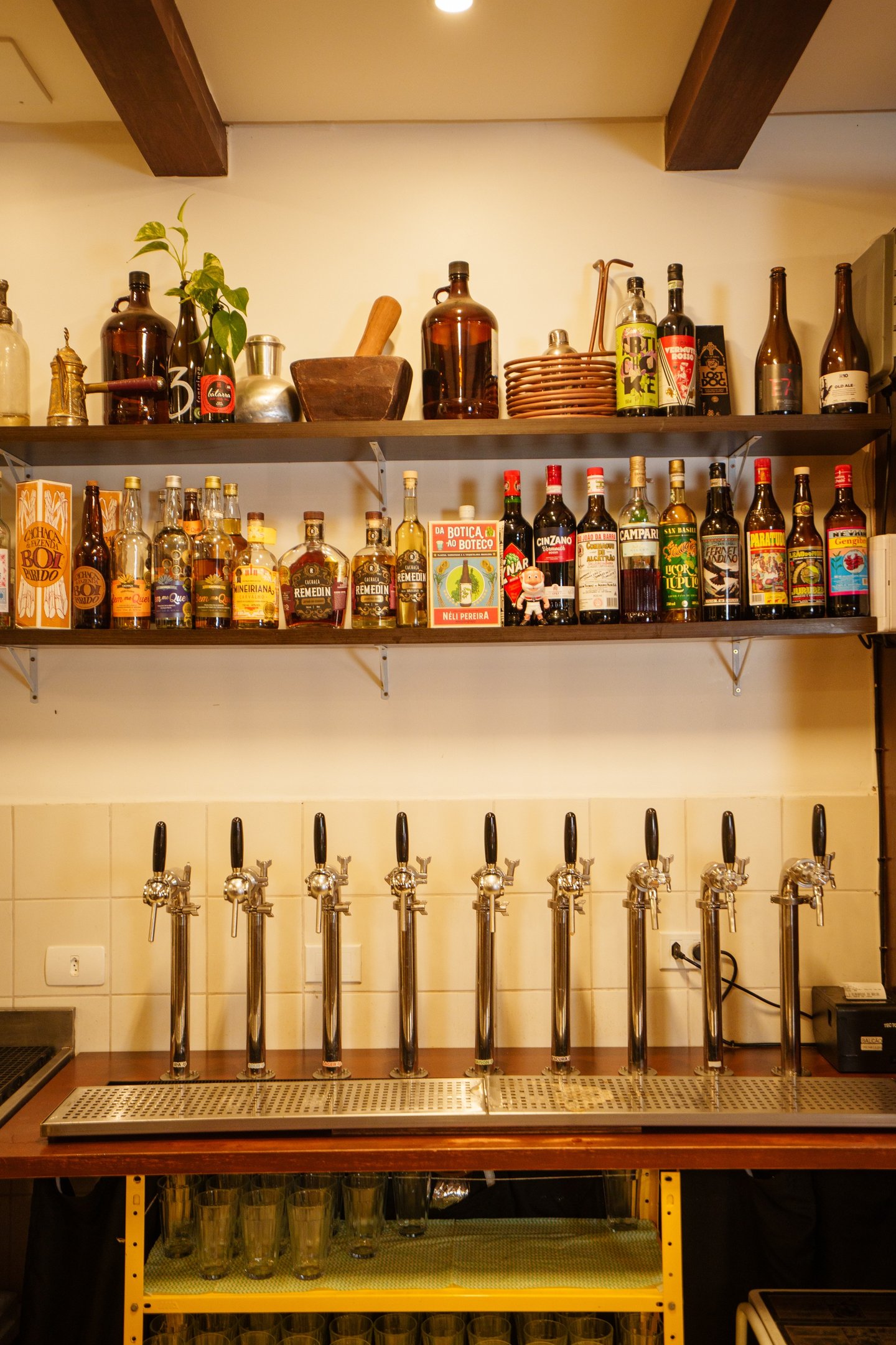 A variety of craft beer tap handles on a bar counter below shelves of liquor bottles and spirits.