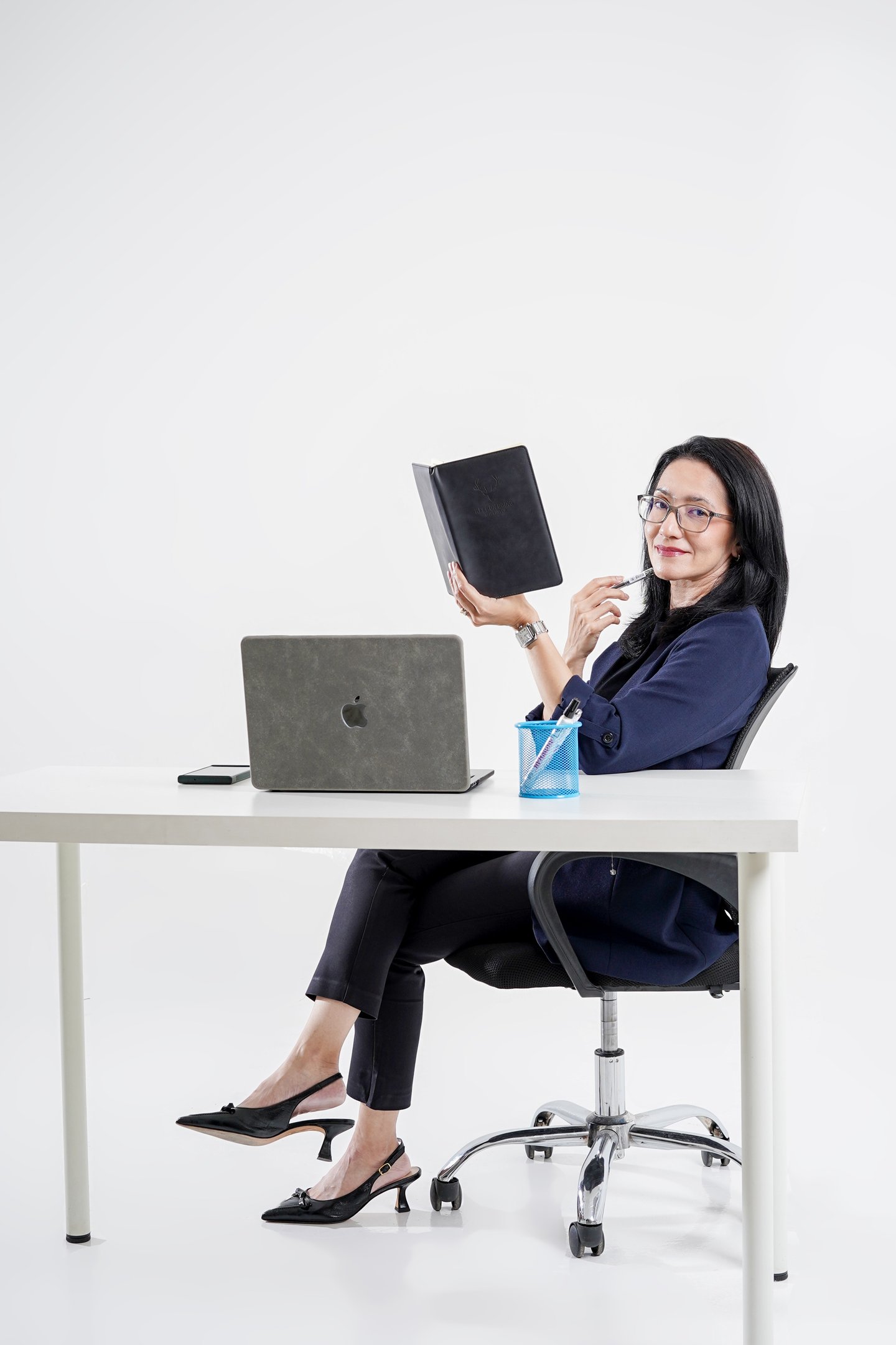 Professional businesswoman in office attire sitting at a desk with a laptop and holding a black notebook.
