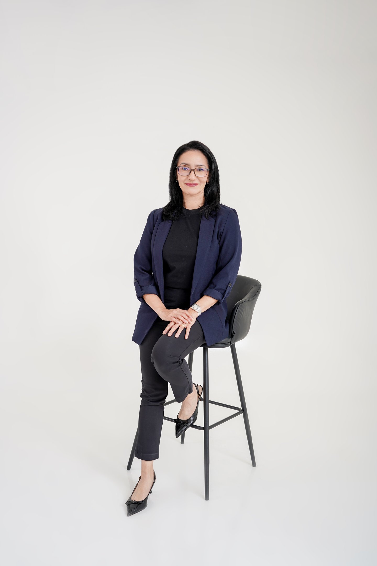 Professional woman in a navy blazer and glasses sitting on a bar stool for a business portrait.