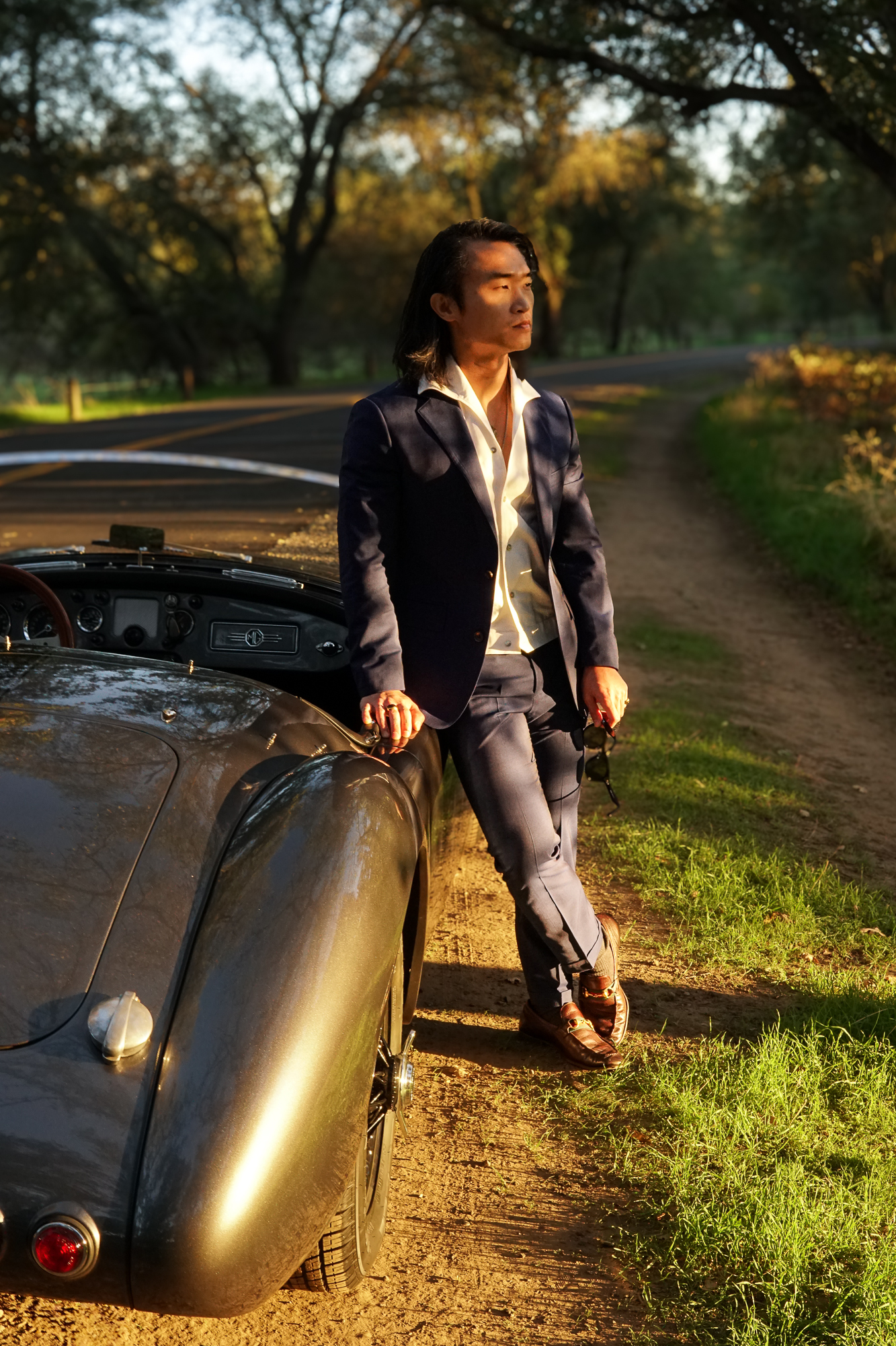 A man leaning against the passenger side of a 1958 MG MGA Roadster in a park during golden hour.
