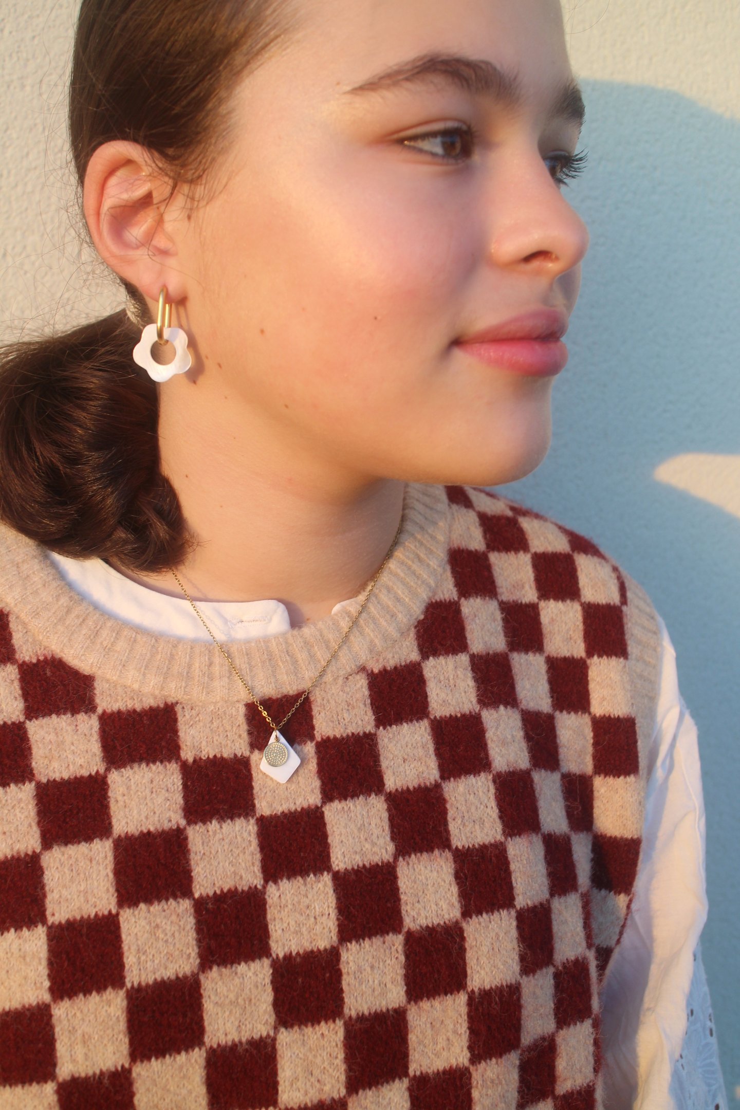 A young woman wearing white floral hoop earrings and a checkered sweater vest in golden hour light.