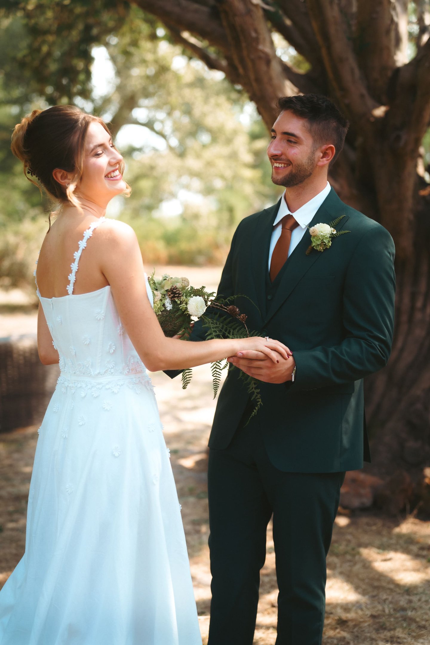 Smiling bride in white floral gown and groom in forest green suit at an outdoor wedding.