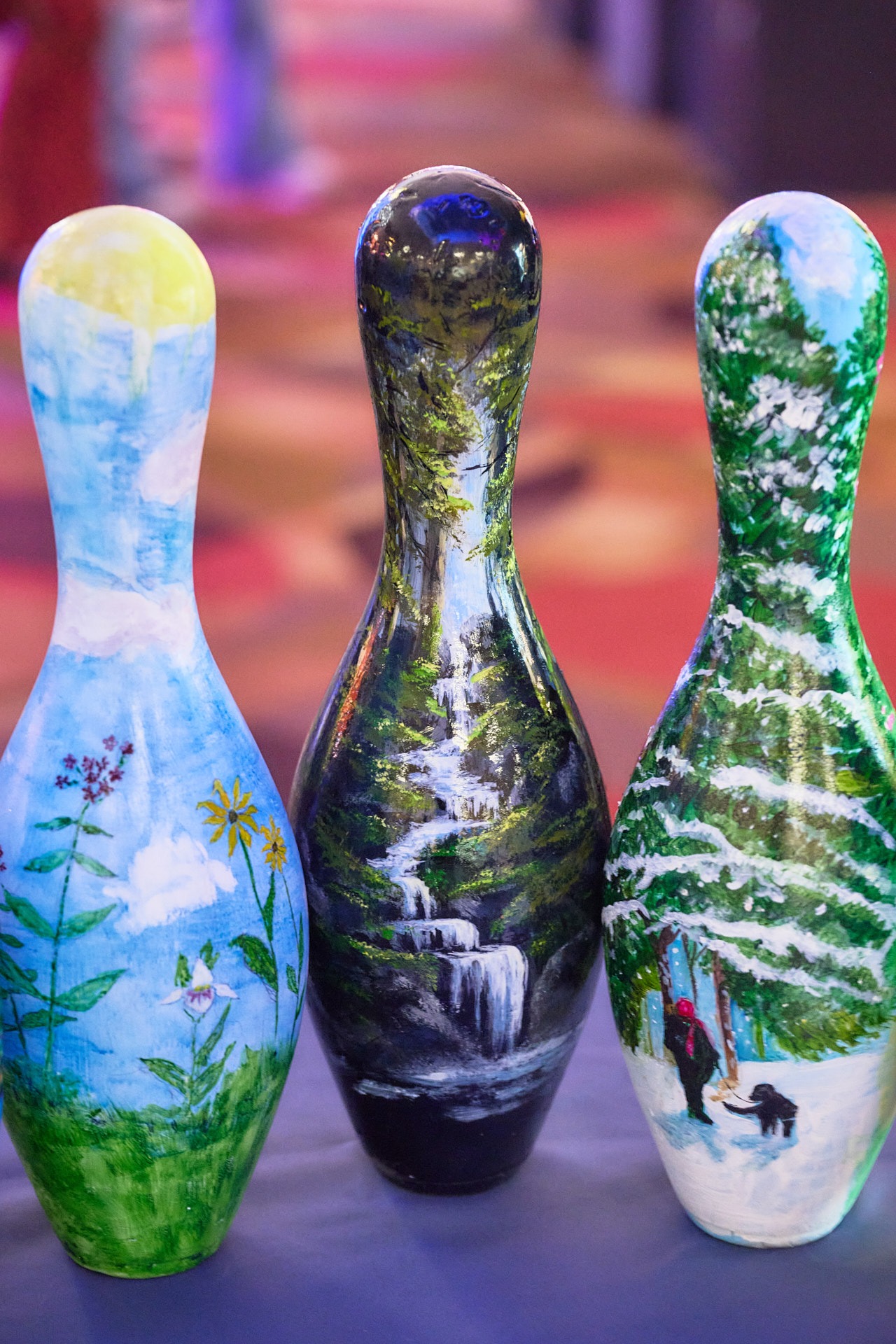 Three hand-painted bowling pins featuring nature landscape art including waterfalls and snowy forests.