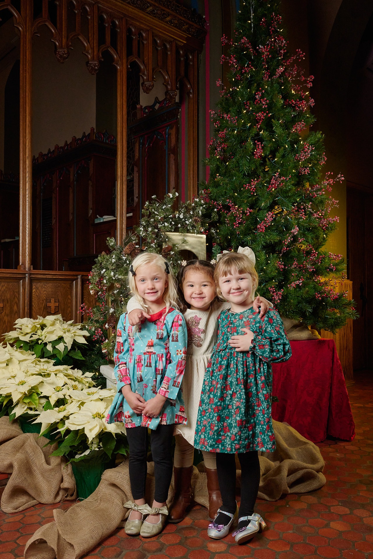 Three young girls in holiday dresses posing by a decorated Christmas tree and white poinsettias.