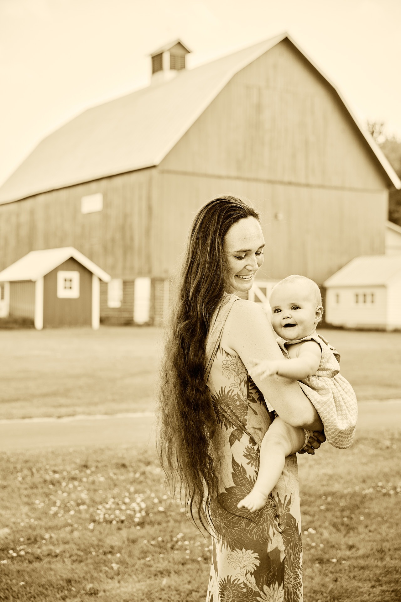 Sepia photograph of a smiling mother holding her happy baby in front of a large rural farm barn.