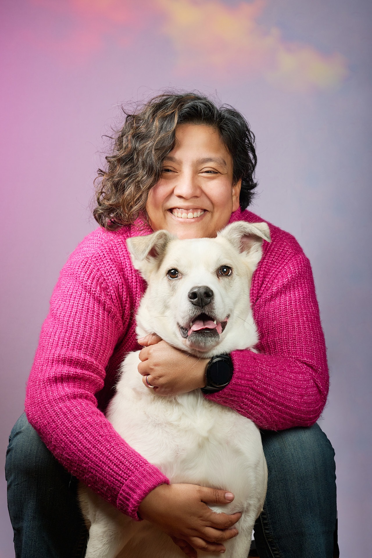 Smiling woman in a pink sweater hugging her large white dog against a purple background.