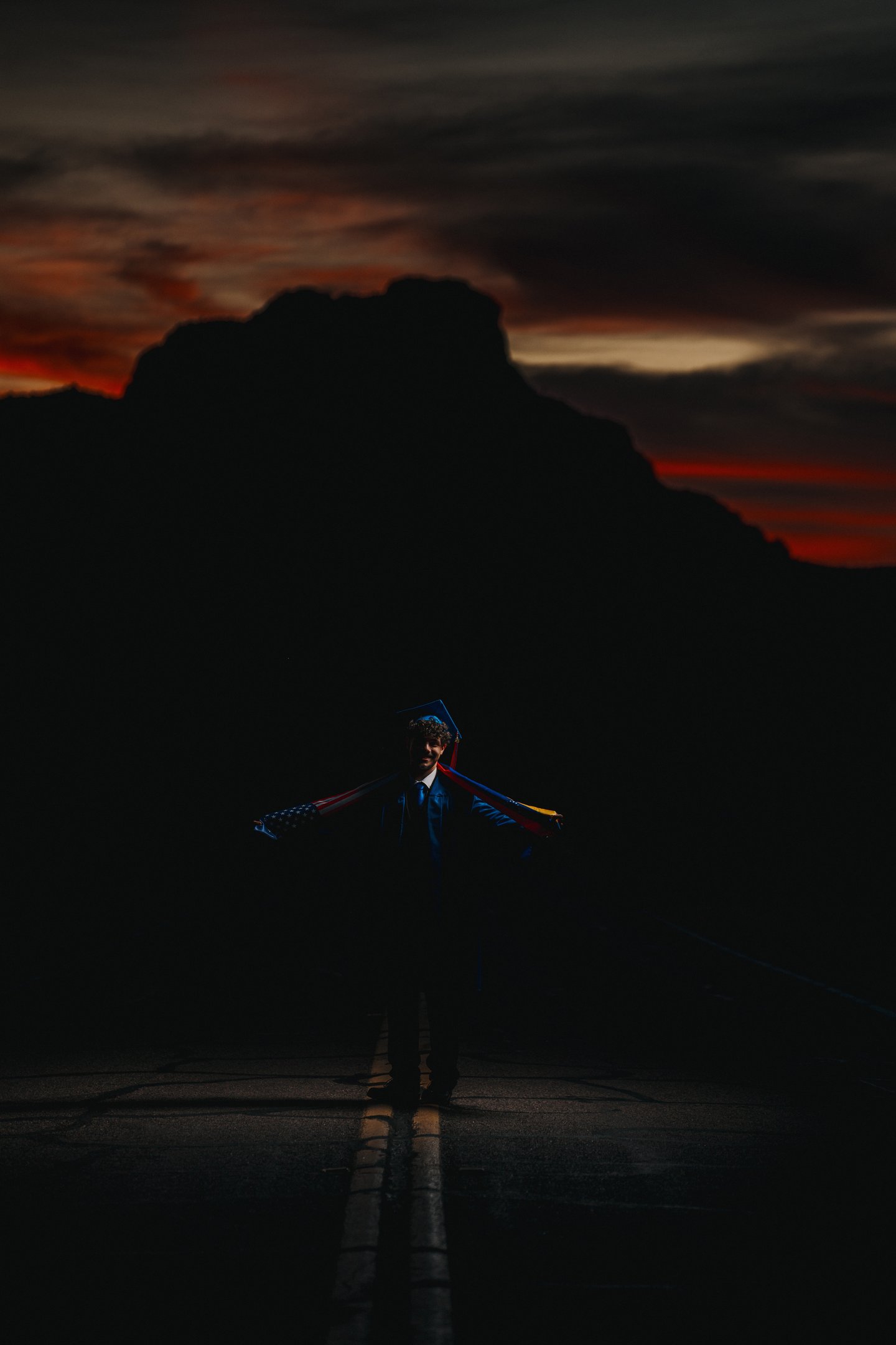 A graduate in cap and gown holds an American flag at sunset with mountain silhouettes.
