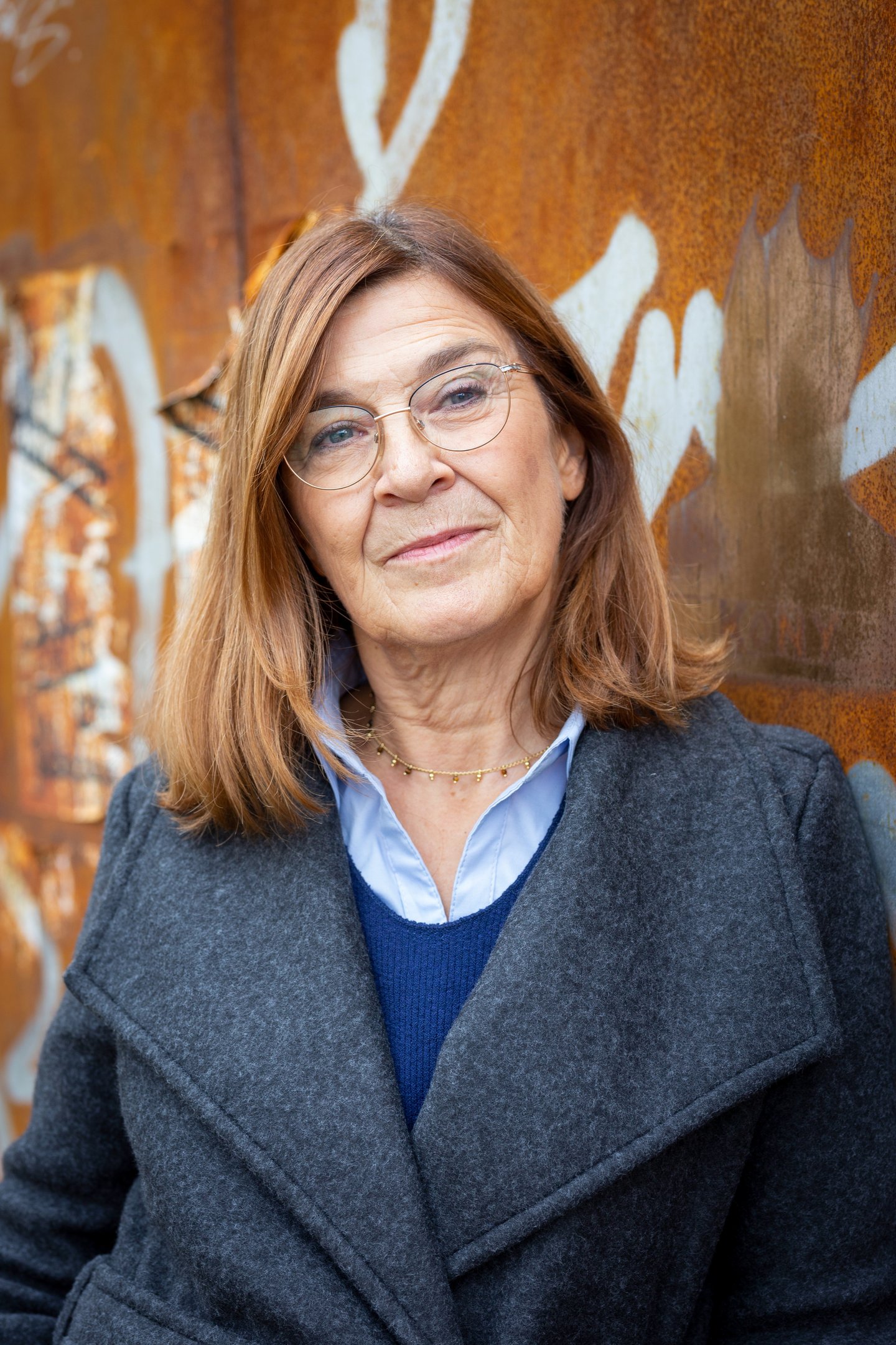 Professional senior woman with glasses and a gray coat standing against a rusty graffiti wall.