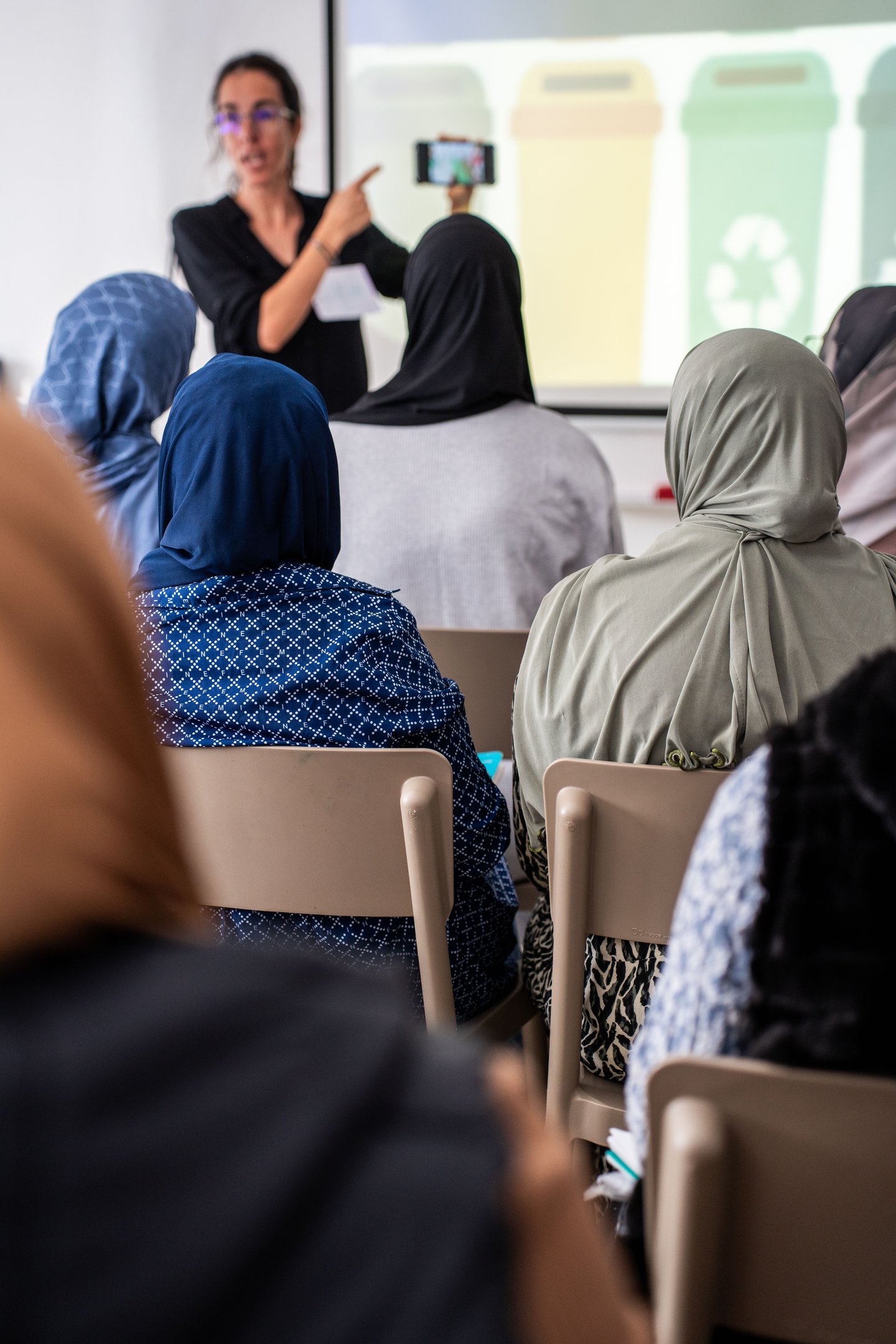 persona blanca con pelo marron y gafas en una aula enseñando un mobil a sus alumnas.