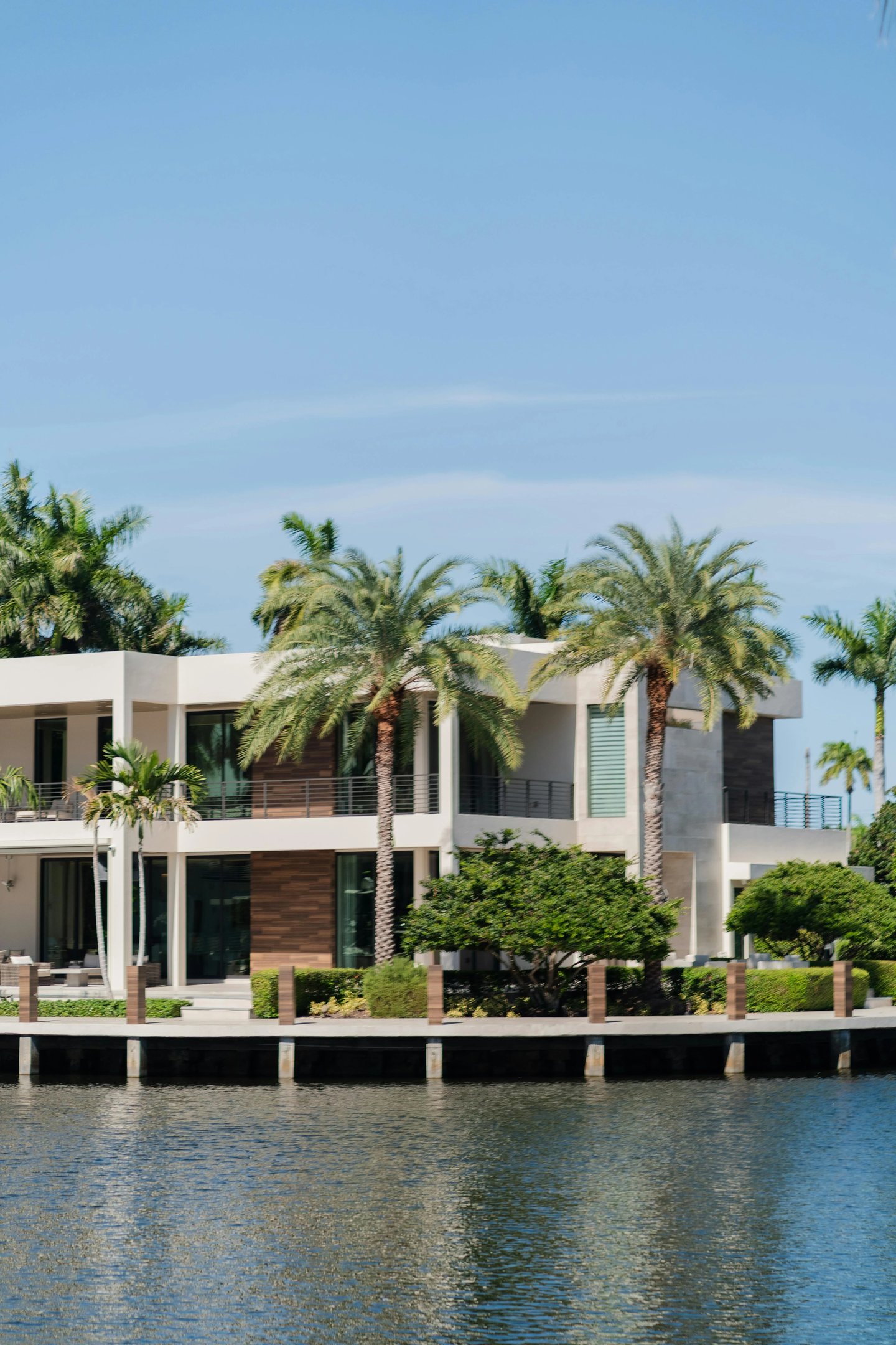 Modern luxury waterfront mansion with palm trees and a private dock on a sunny day.