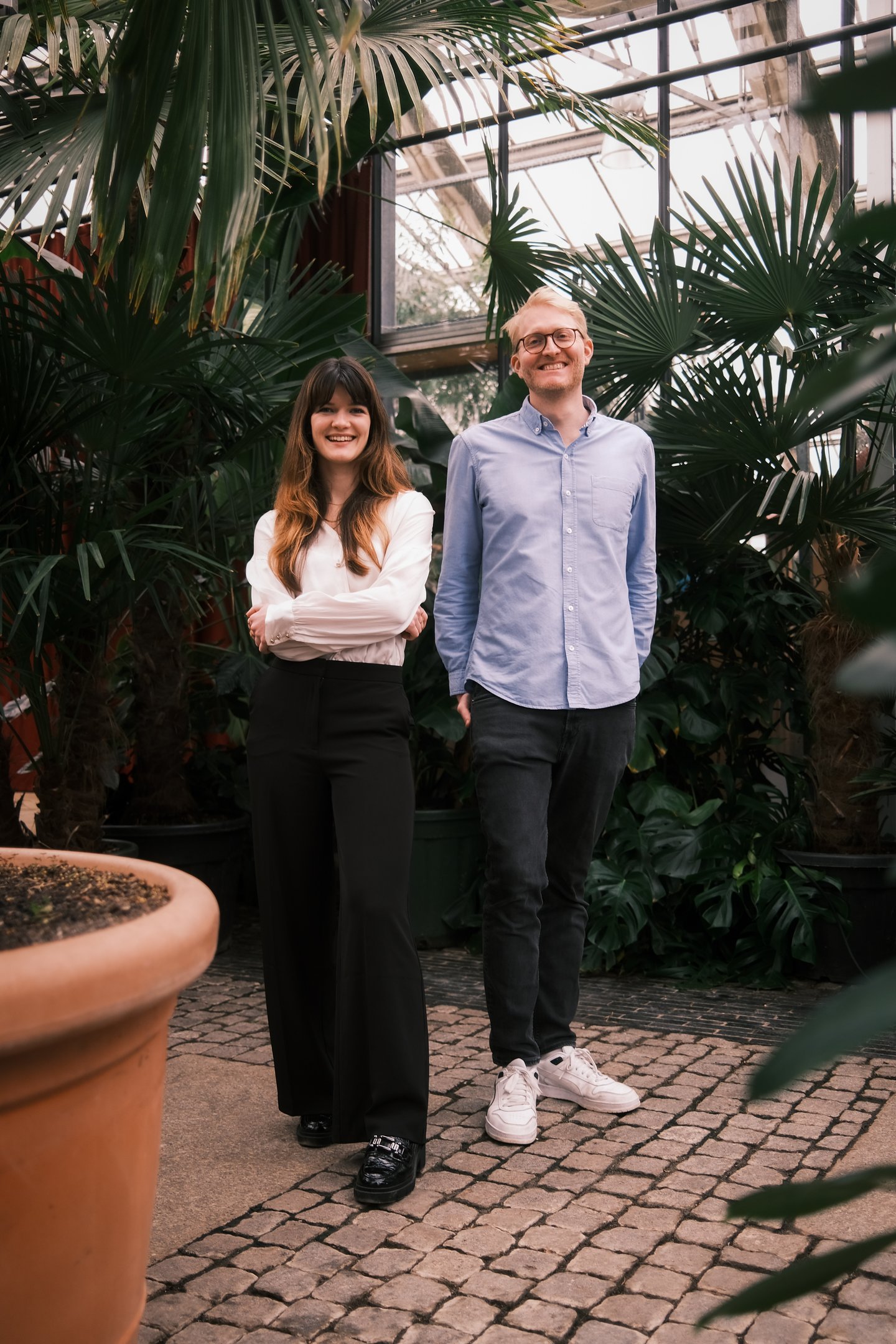 A smiling man and woman standing together in a lush indoor greenhouse surrounded by tropical plants.