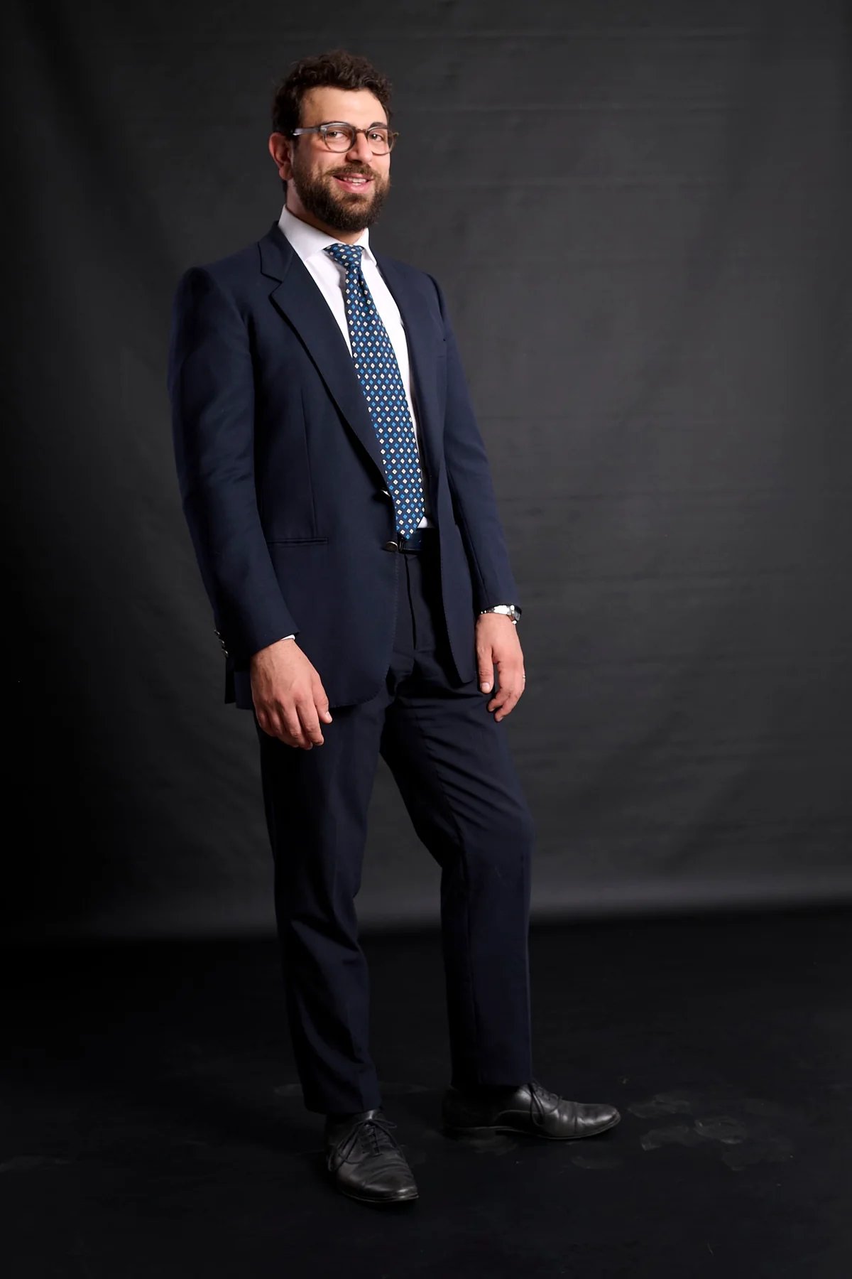 Professional man in a navy blue suit and patterned tie posing for a corporate headshot against a black background.