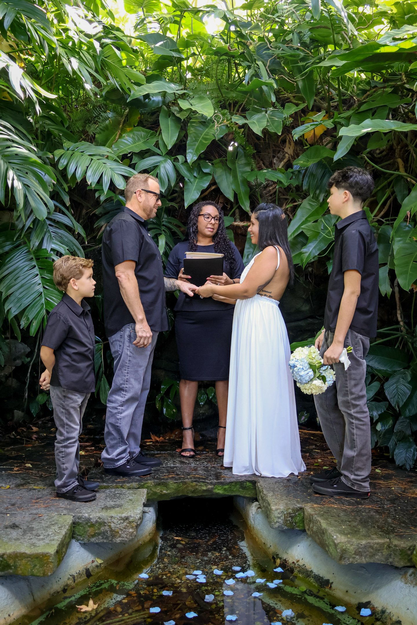 A couple exchanges vows at an outdoor tropical garden wedding ceremony with an officiant and children.