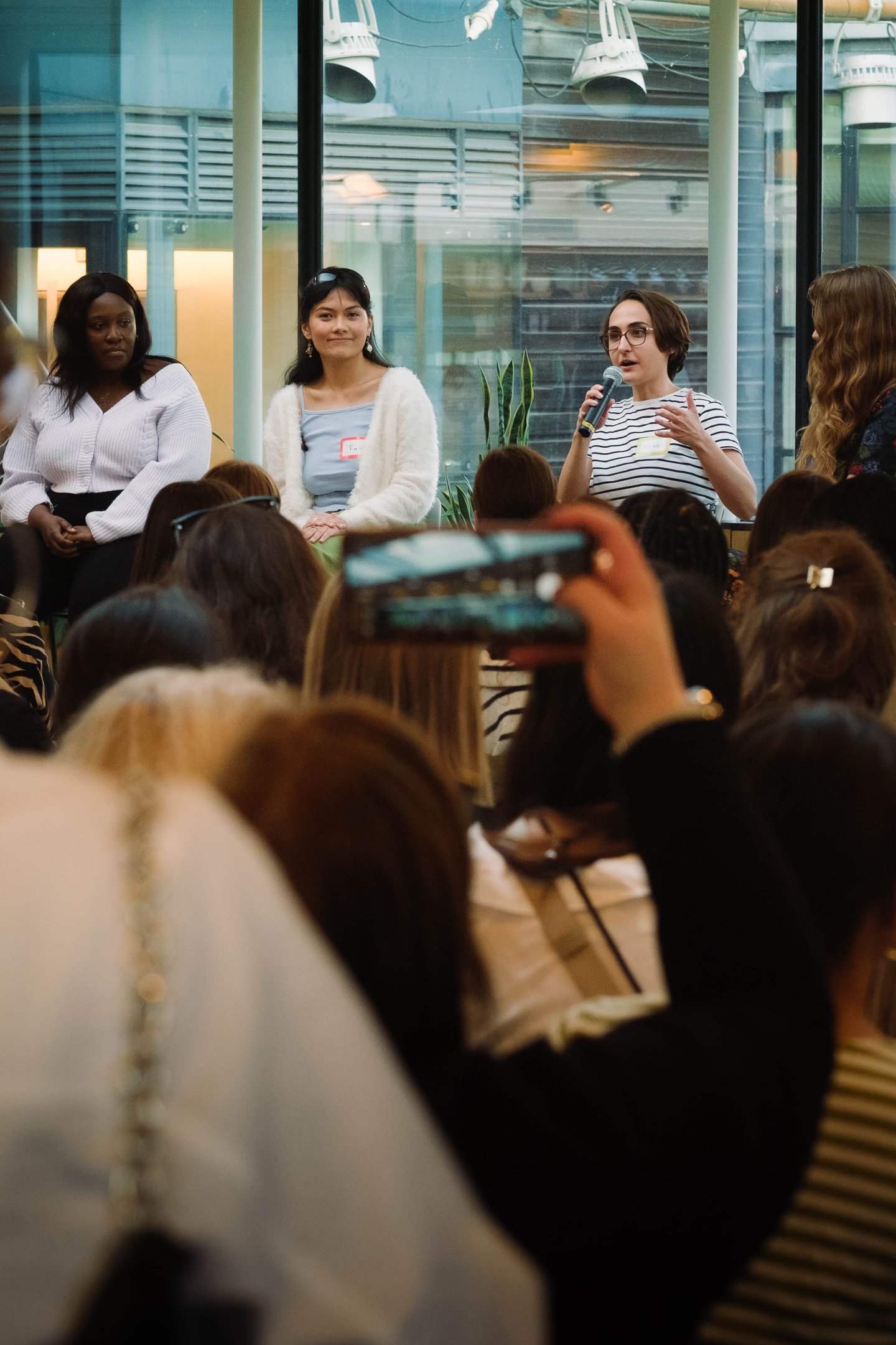 Photo of Micha in a stripy t-shirt holding a microphone and speaking to an audience