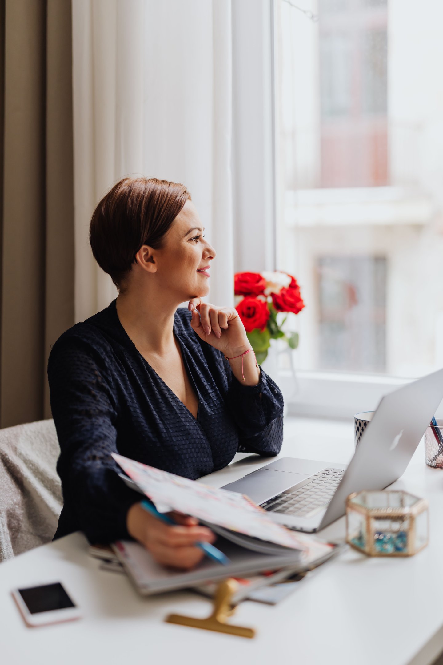 Professional female entrepreneur working on her laptop at a home office desk by a window.