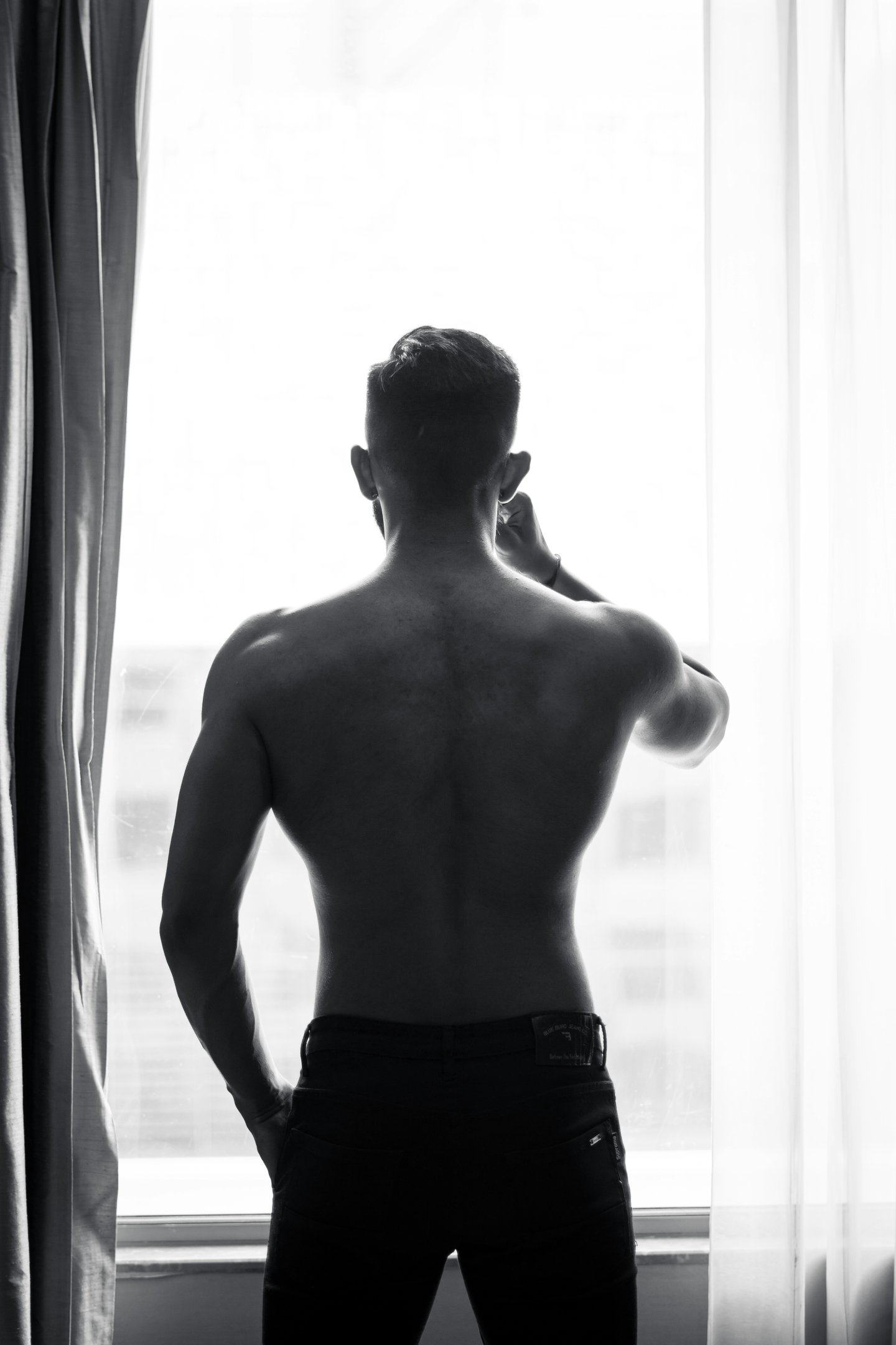 Silhouette of a muscular shirtless man looking out a bright window in black and white.