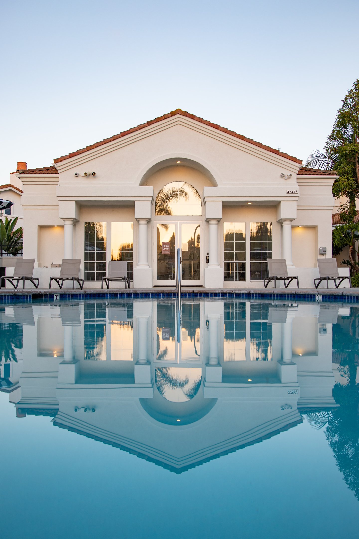 Off-white facade of luxury building with pillar architecture. A pool in front and deckchairs.