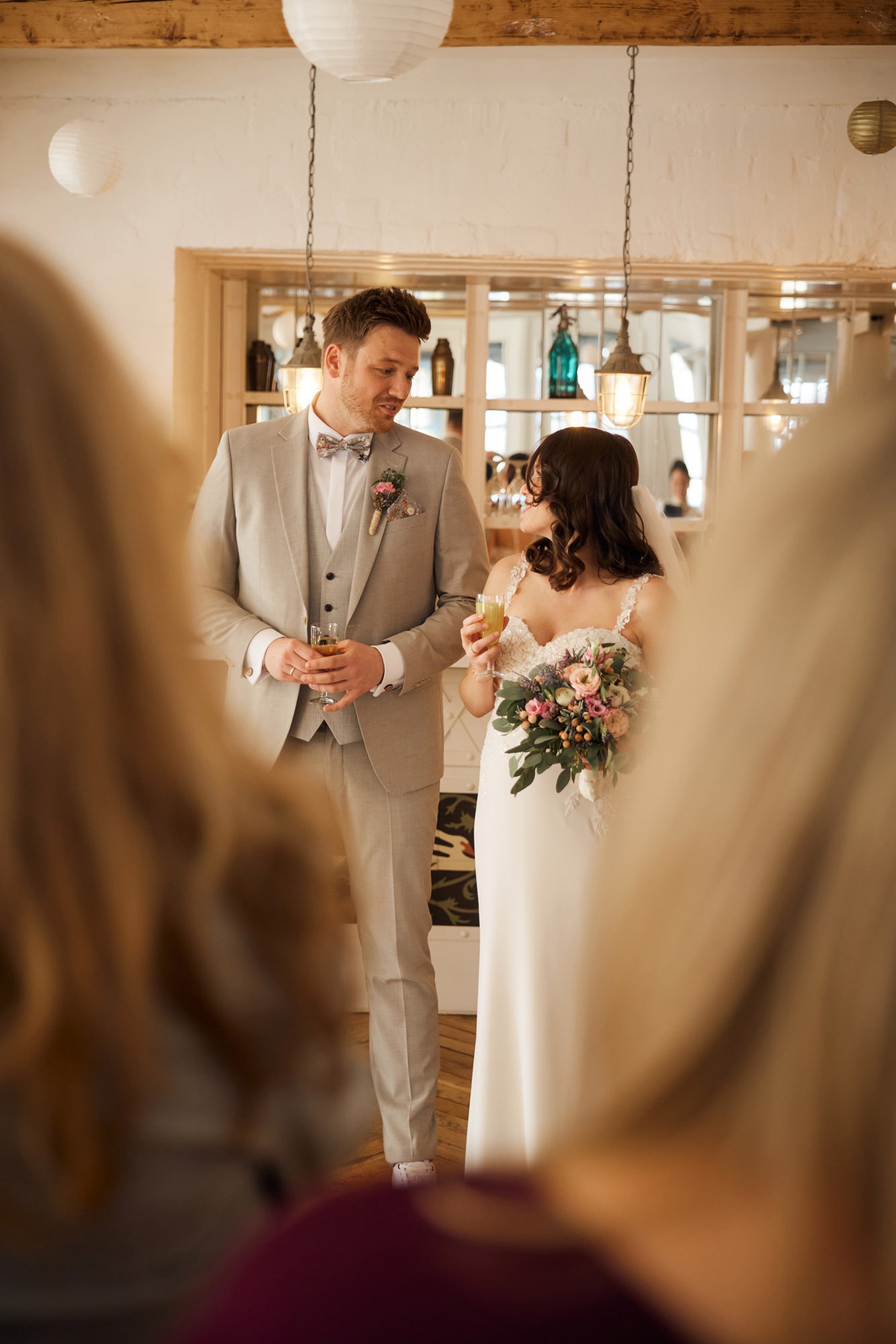 A bride and groom in a light grey suit holding drinks and flowers at their indoor wedding reception.