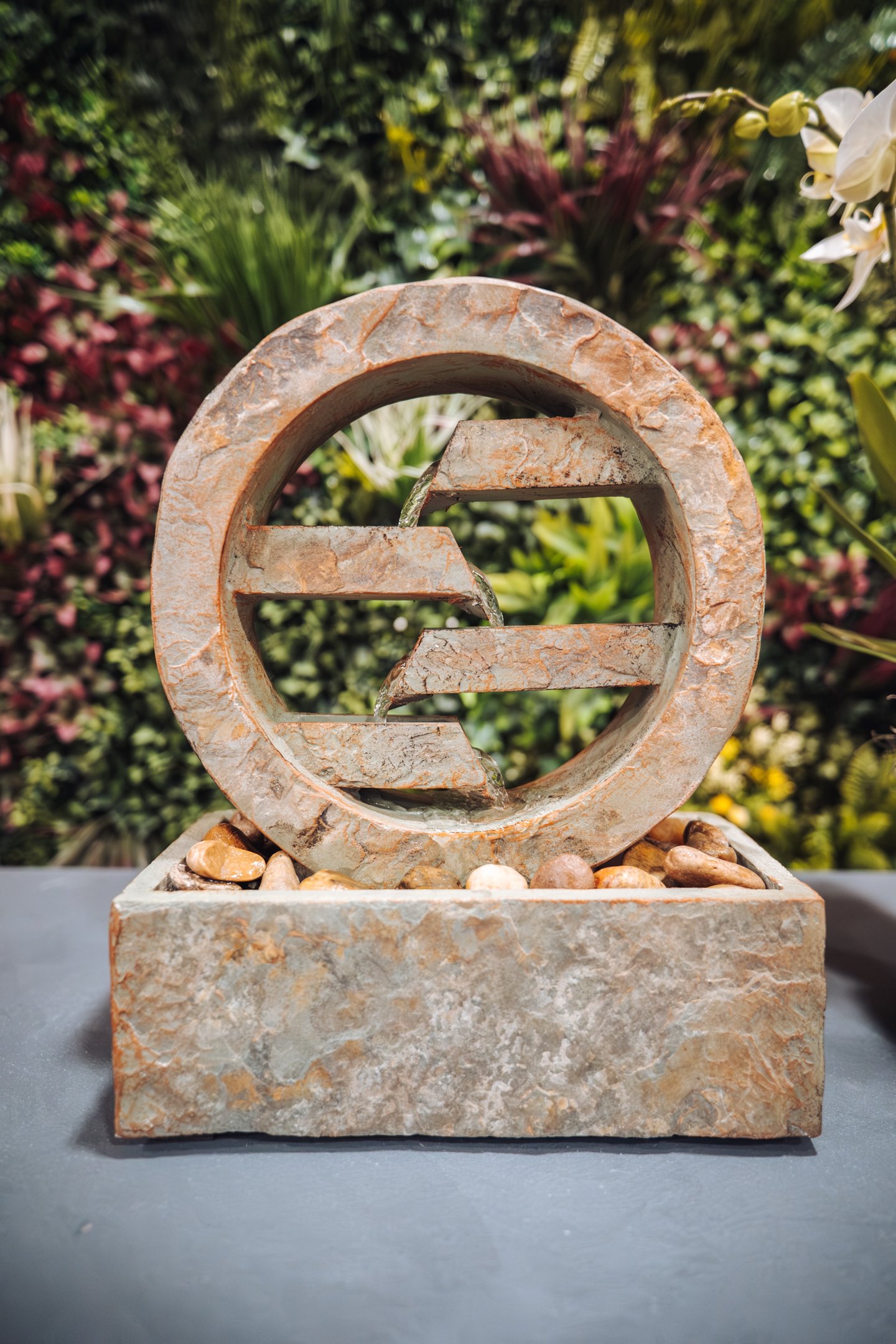 A circular stone tiered desktop water fountain with flowing water against a lush green wall.