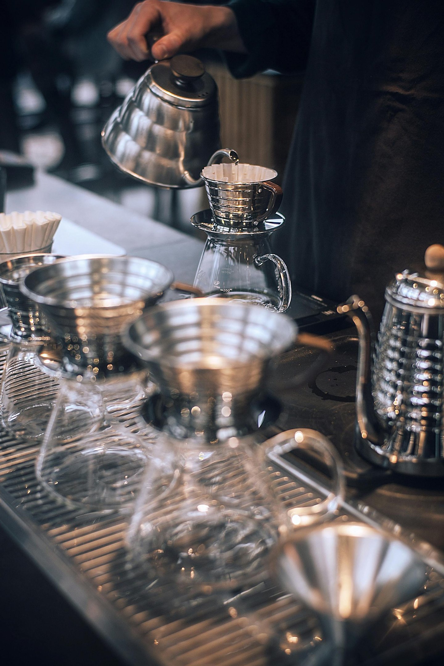 Barista pouring hot water from a gooseneck kettle into a stainless steel pour-over coffee dripper.