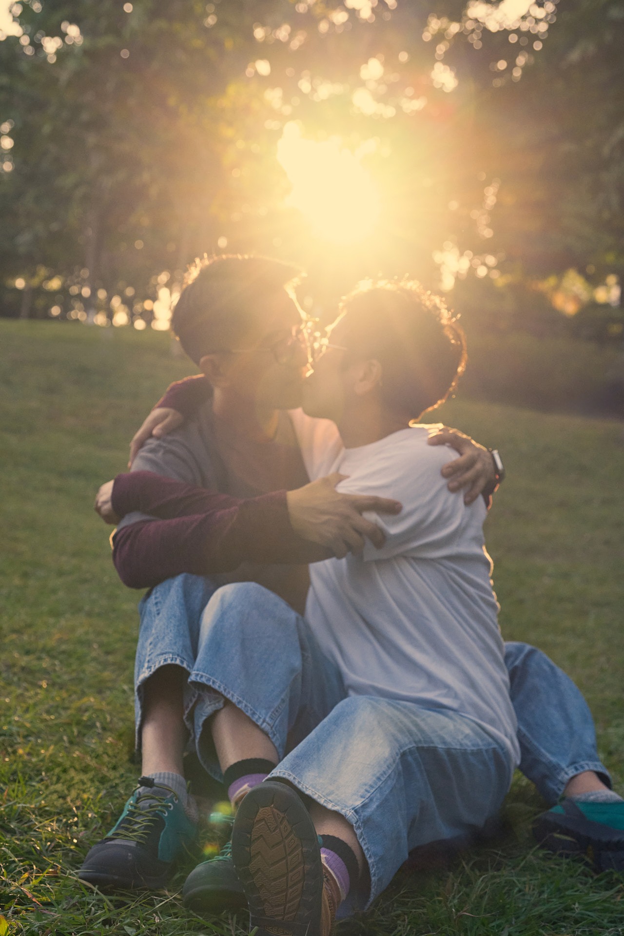 A couple kissing while sitting on the grass in a park during a golden hour sunset.